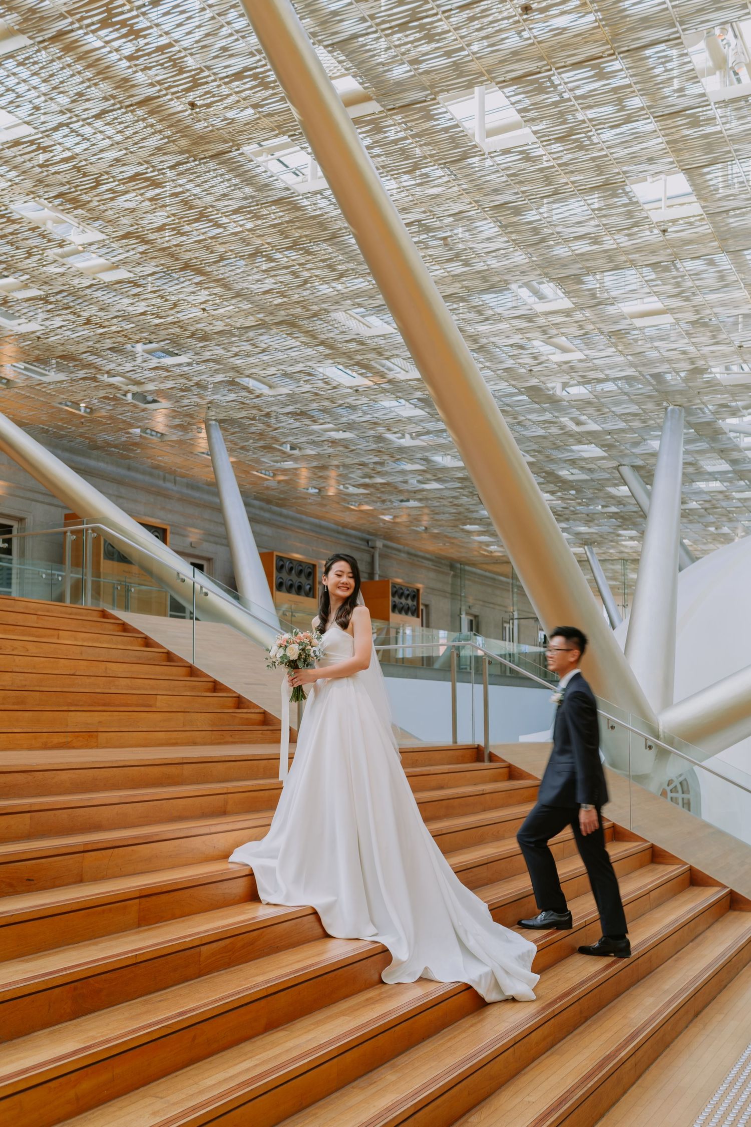 Bride and groom posing on grand wooden stairs beneath a stunning latticed ceiling.