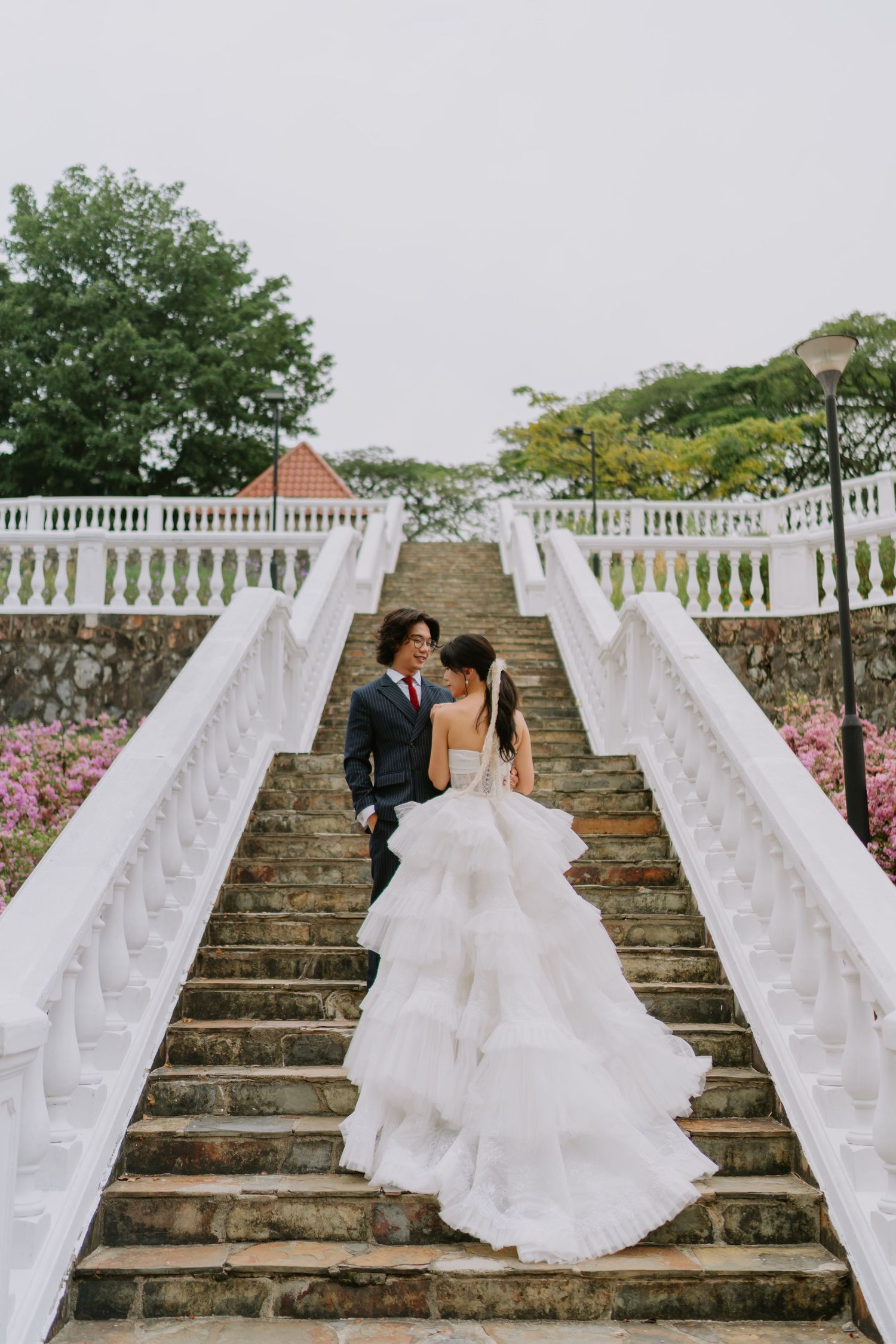 Bride and groom facing each other on grand stone steps flanked by white railings and pink flowering shrubs.