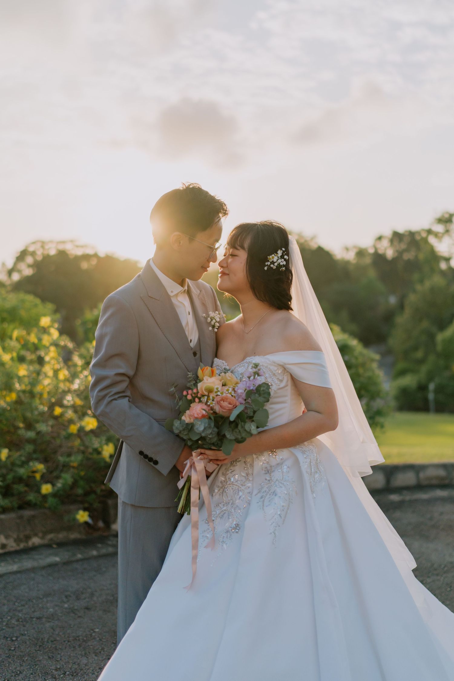 Romantic couple nearly kissing at sunset, bride in off-shoulder beaded gown holding colorful bouquet.