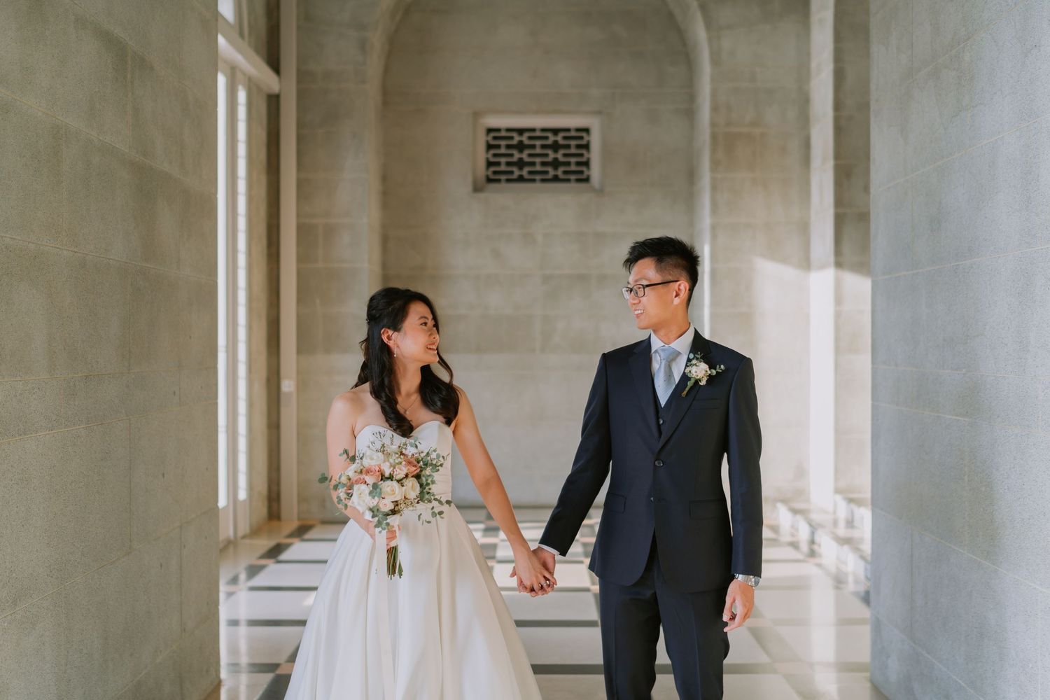 Bride and groom hold hands smiling in sunlit stone colonnade during wedding photoshoot.