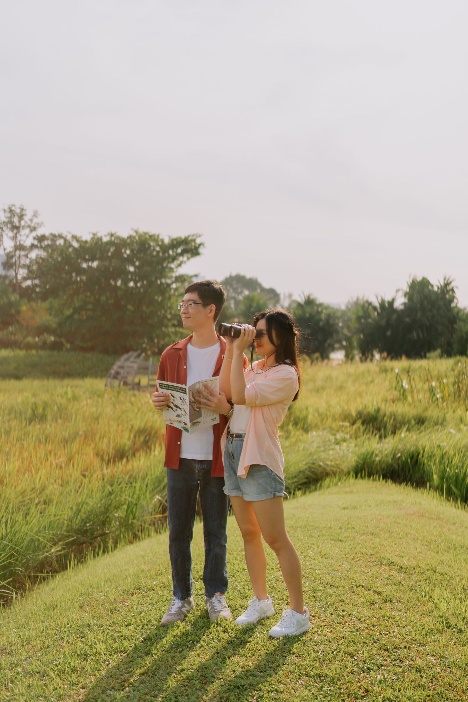 A couple explores nature, with the woman using binoculars and the man holding a map outdoors.
