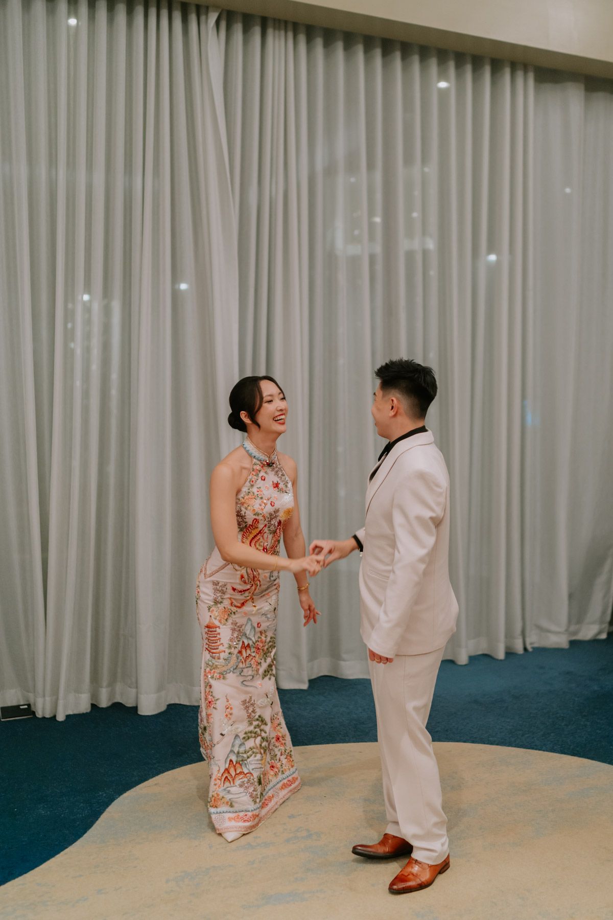 Couple in elegant attire holding hands, woman in floral cheongsam and man in white suit, standing before gray curtains.