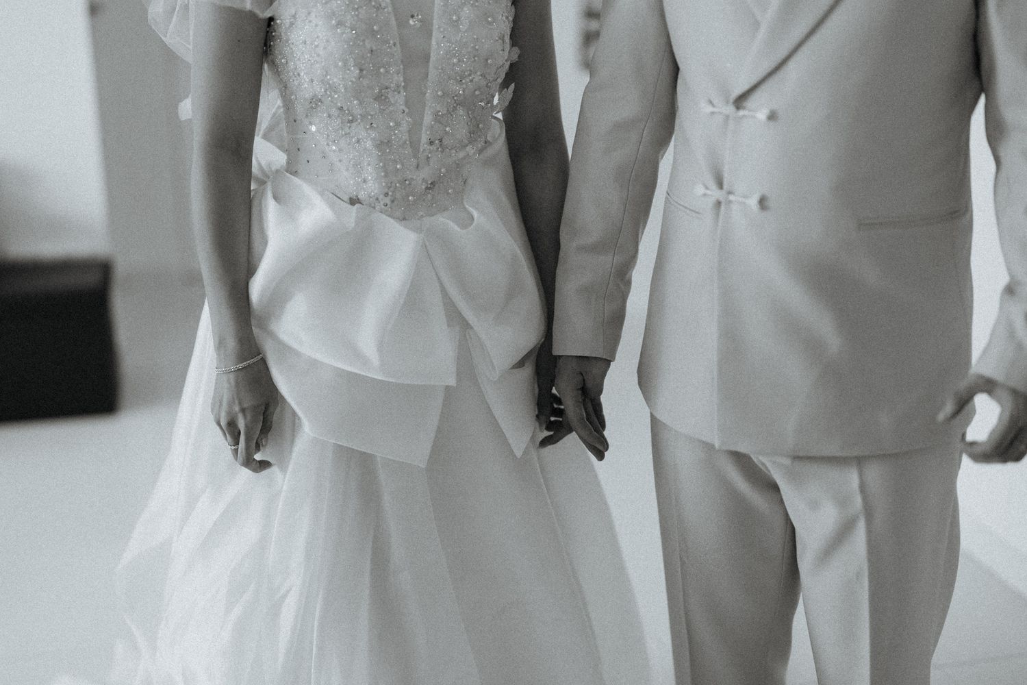 Black and white photo of a bride and groom holding hands, wearing elegant wedding attire.