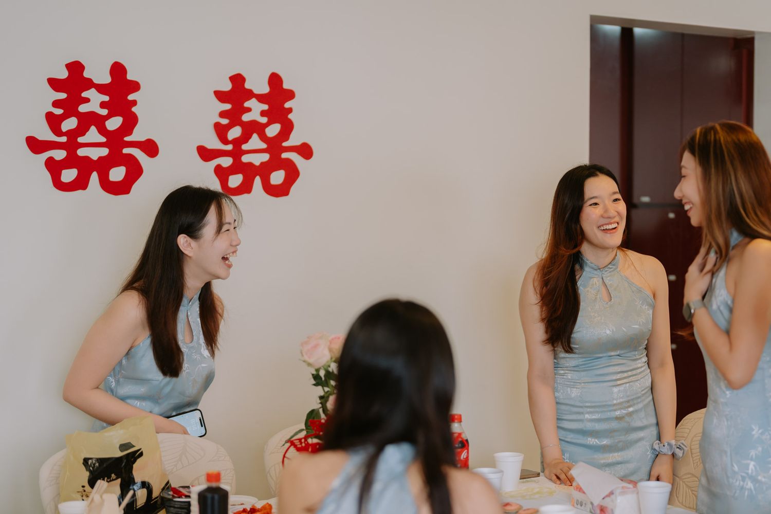 Women in blue dresses laugh together at a Chinese wedding celebration with double happiness symbols on the wall.
