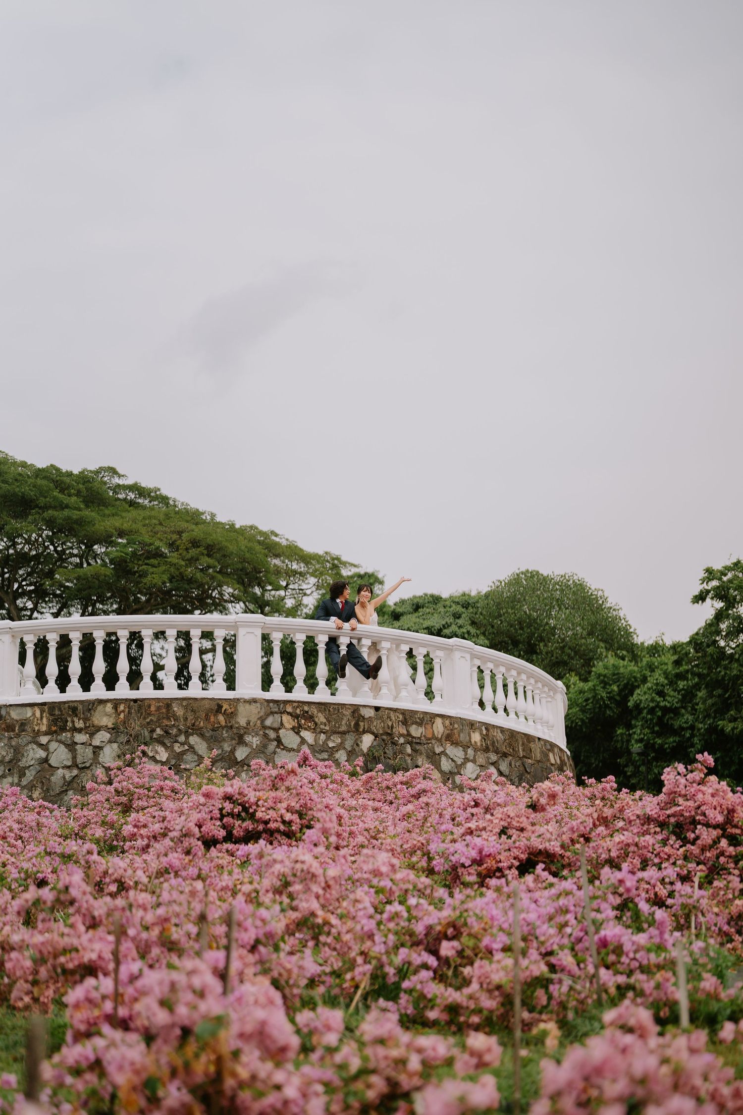 Bride and groom seated on a curved white stone balcony overlooking a garden of blooming pink flowers.