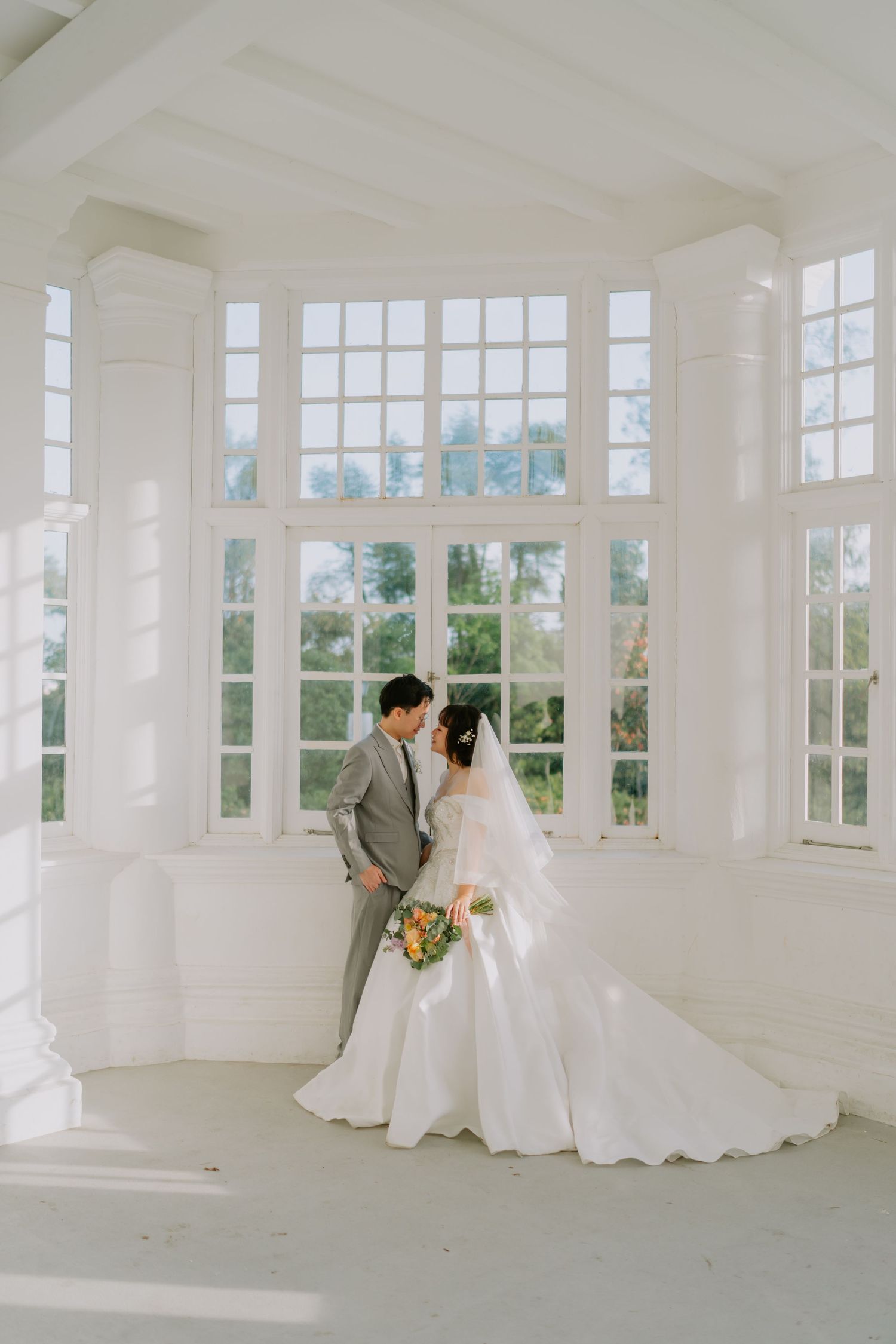 Bride and groom stand together in a sunlit white room with large arched windows and lush greenery outside.