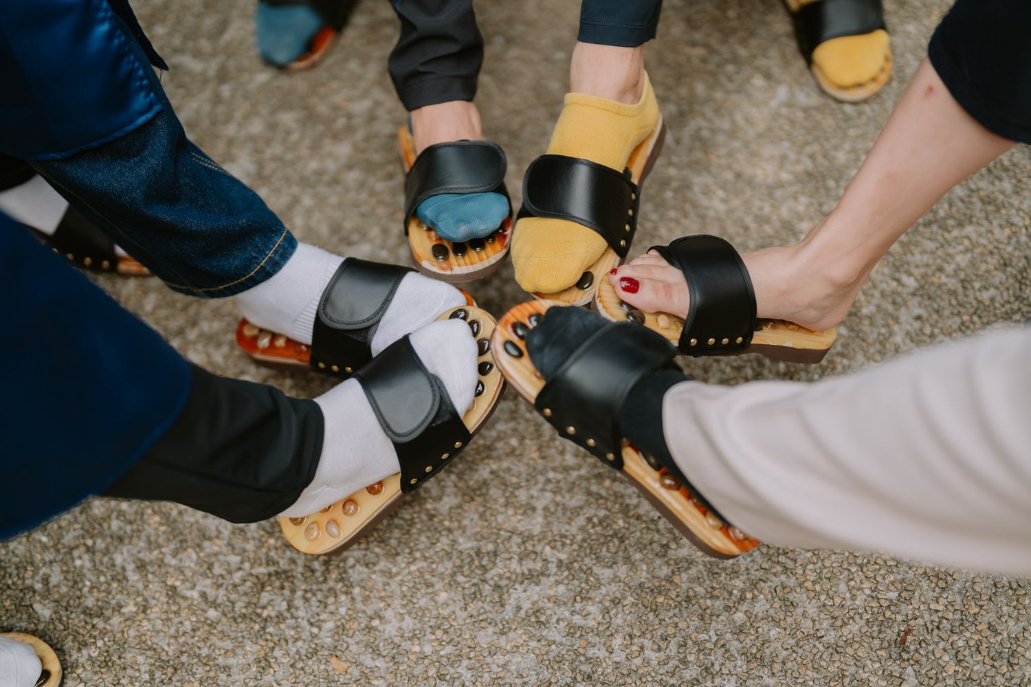 Group of people wearing stylish black and tan wooden clogs and sandals arranged in a circle on gray pavement.