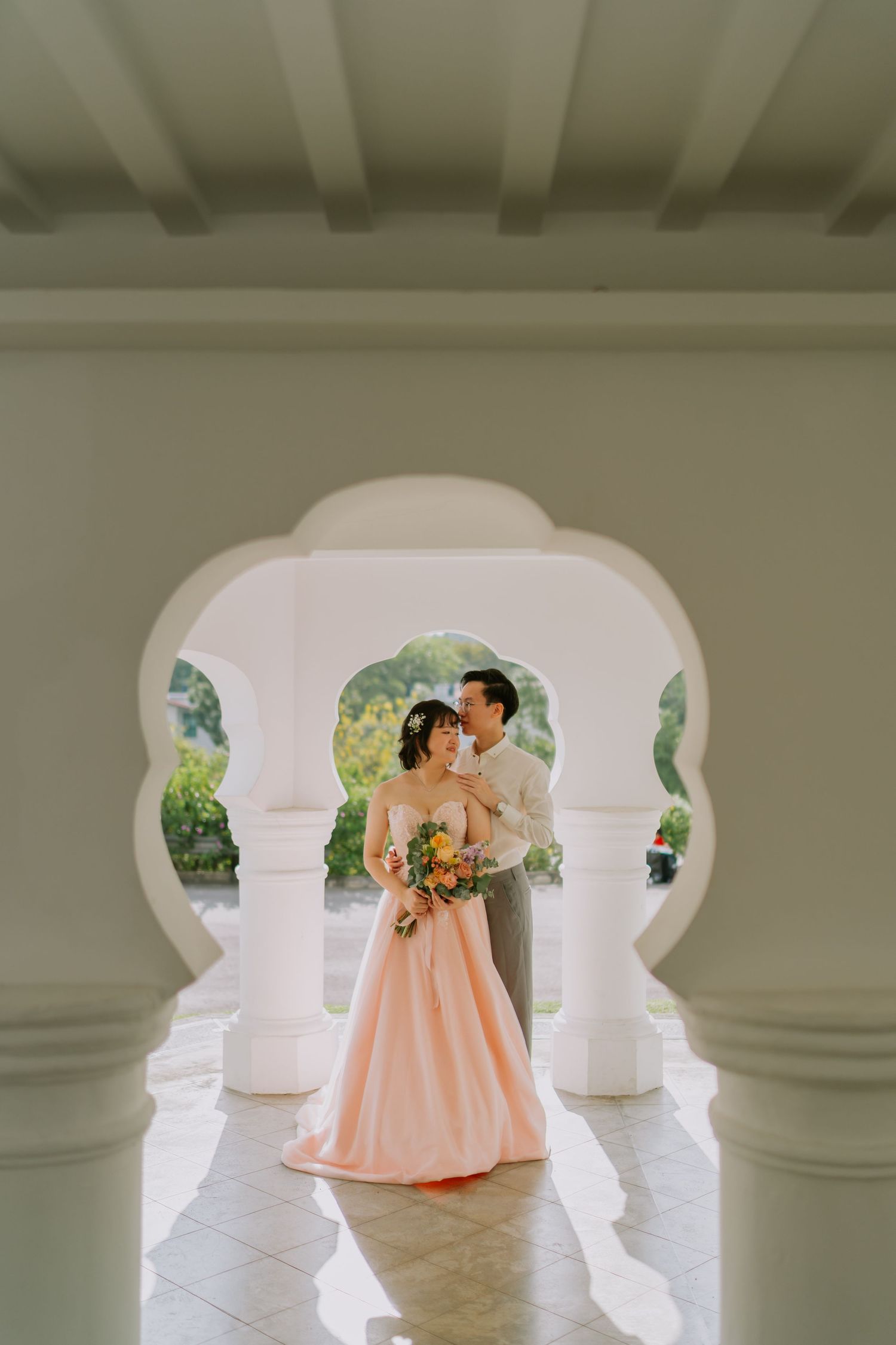 Bride in pink gown and groom embracing under elegant white Moorish arches with lush greenery.