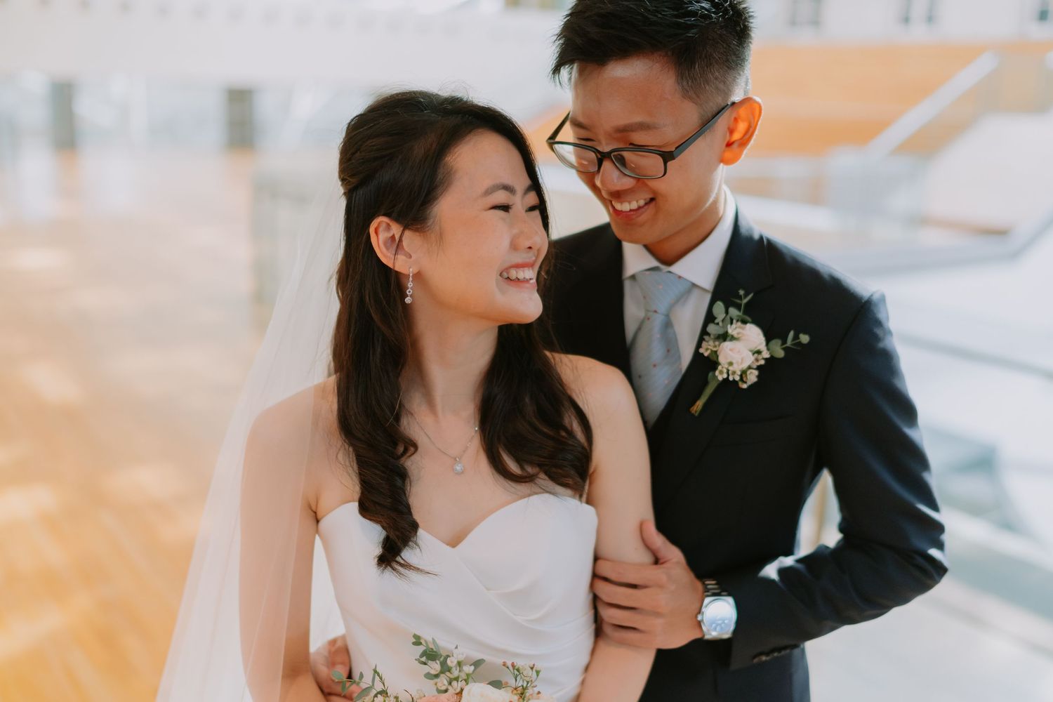 Smiling Asian bride in strapless white gown with groom in navy suit sharing joyful outdoor wedding moment.