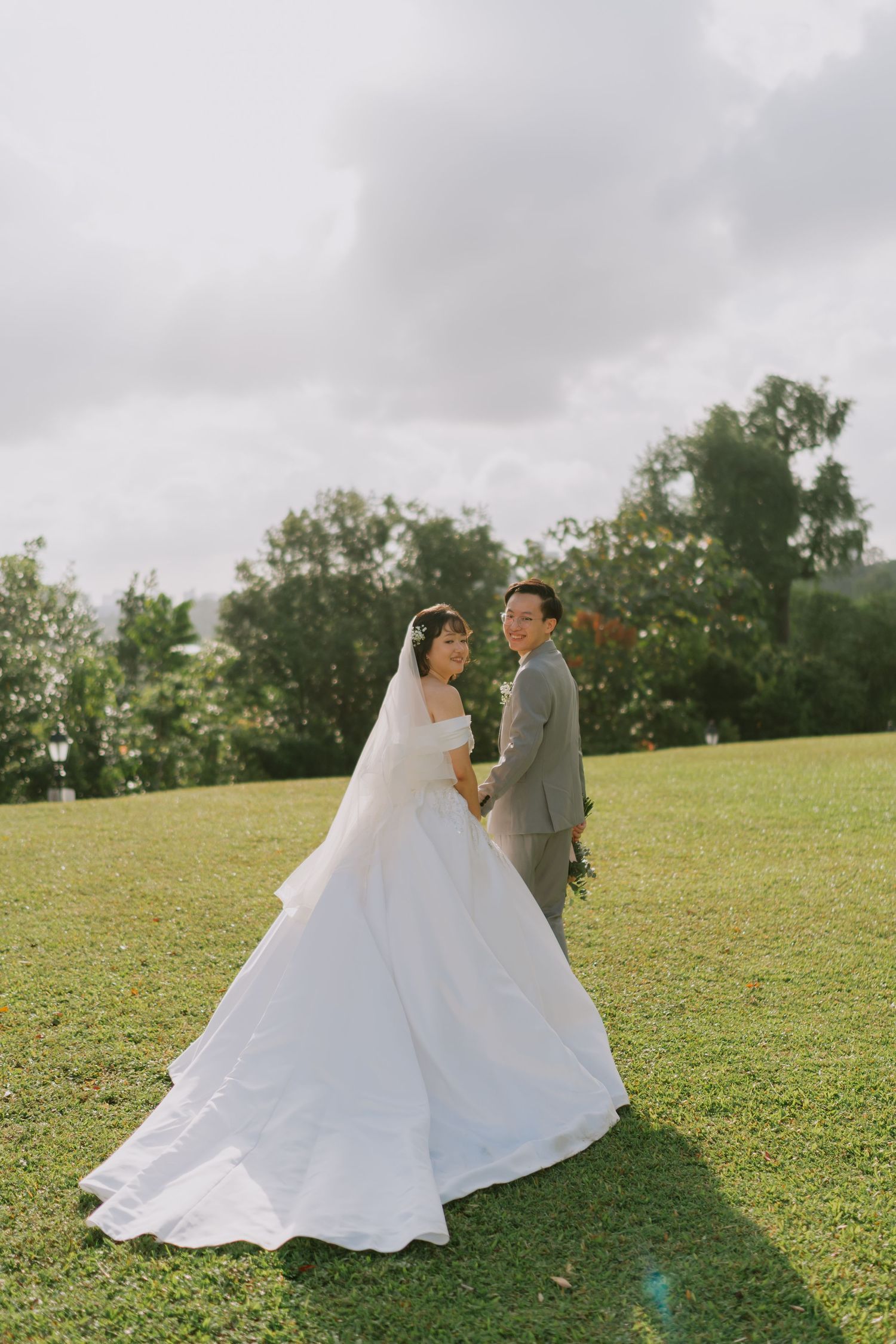 Bride in a flowing white gown and groom in gray suit pose on a green hillside lawn.