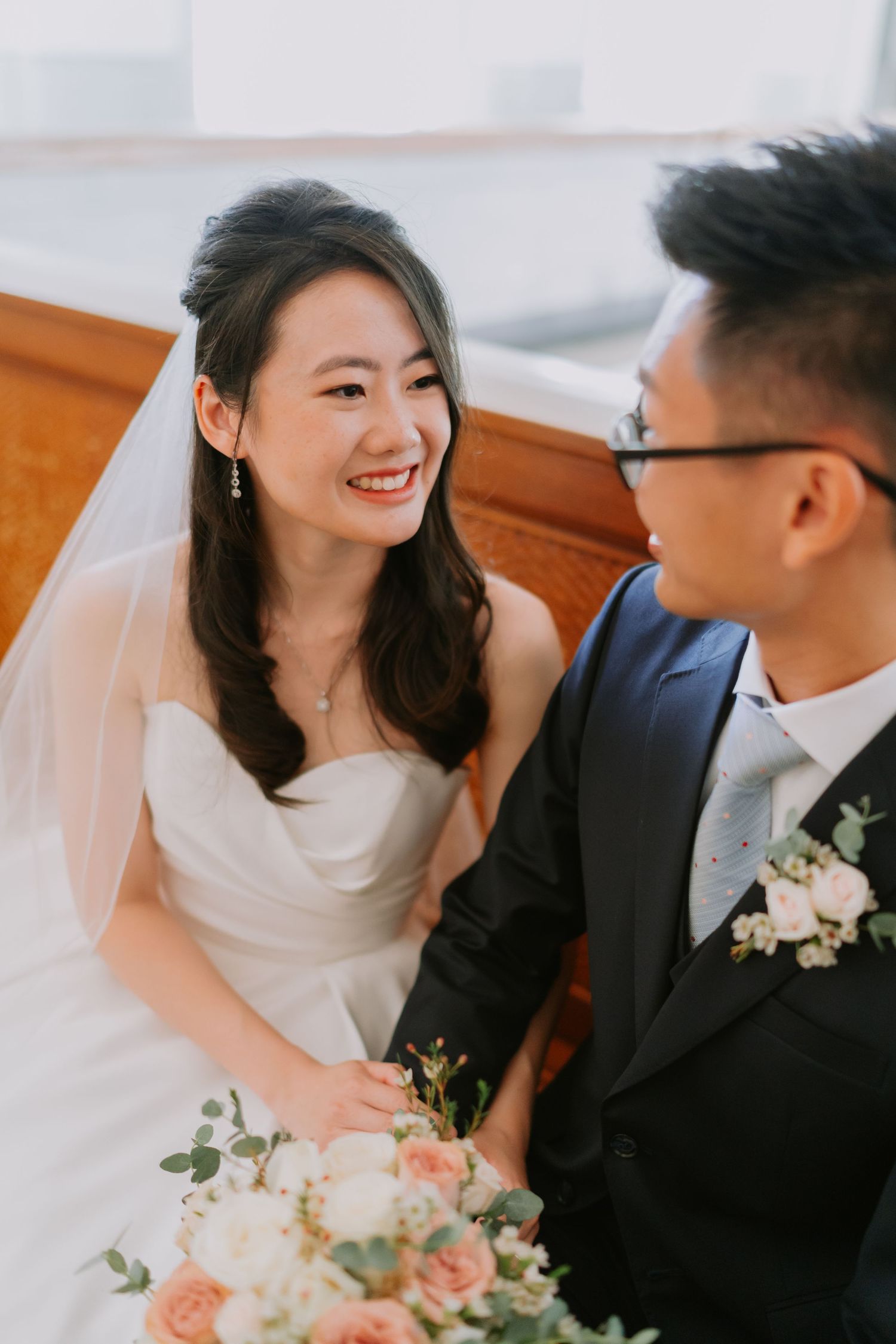 Smiling Asian bride in white strapless gown with veil laughing joyfully with groom in dark suit at church.