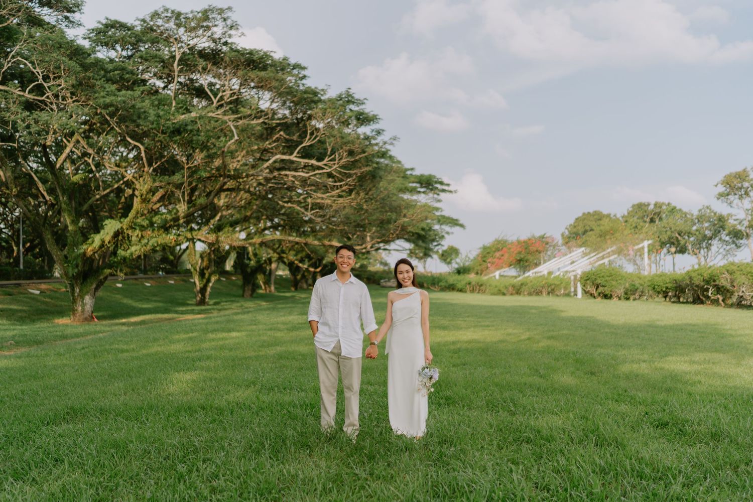 Asian wedding couple holding hands while walking across lush green lawn under blue sky.