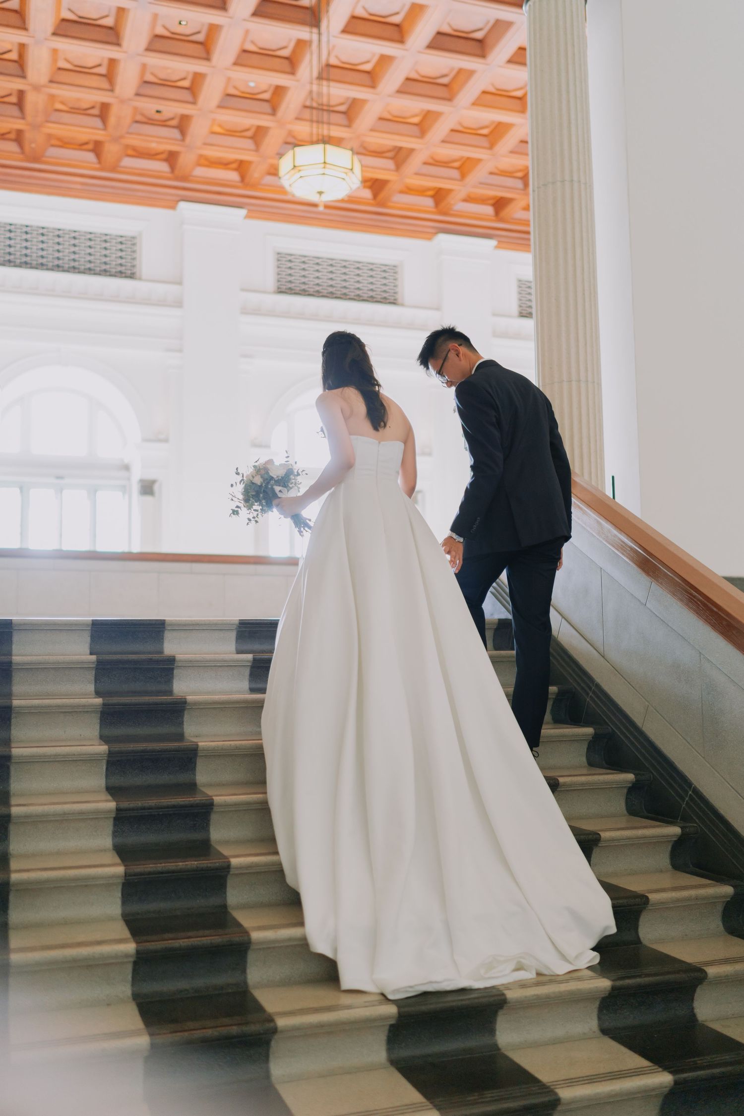 Bride and groom walking up grand staircase hand in hand, bride in elegant white ballgown.