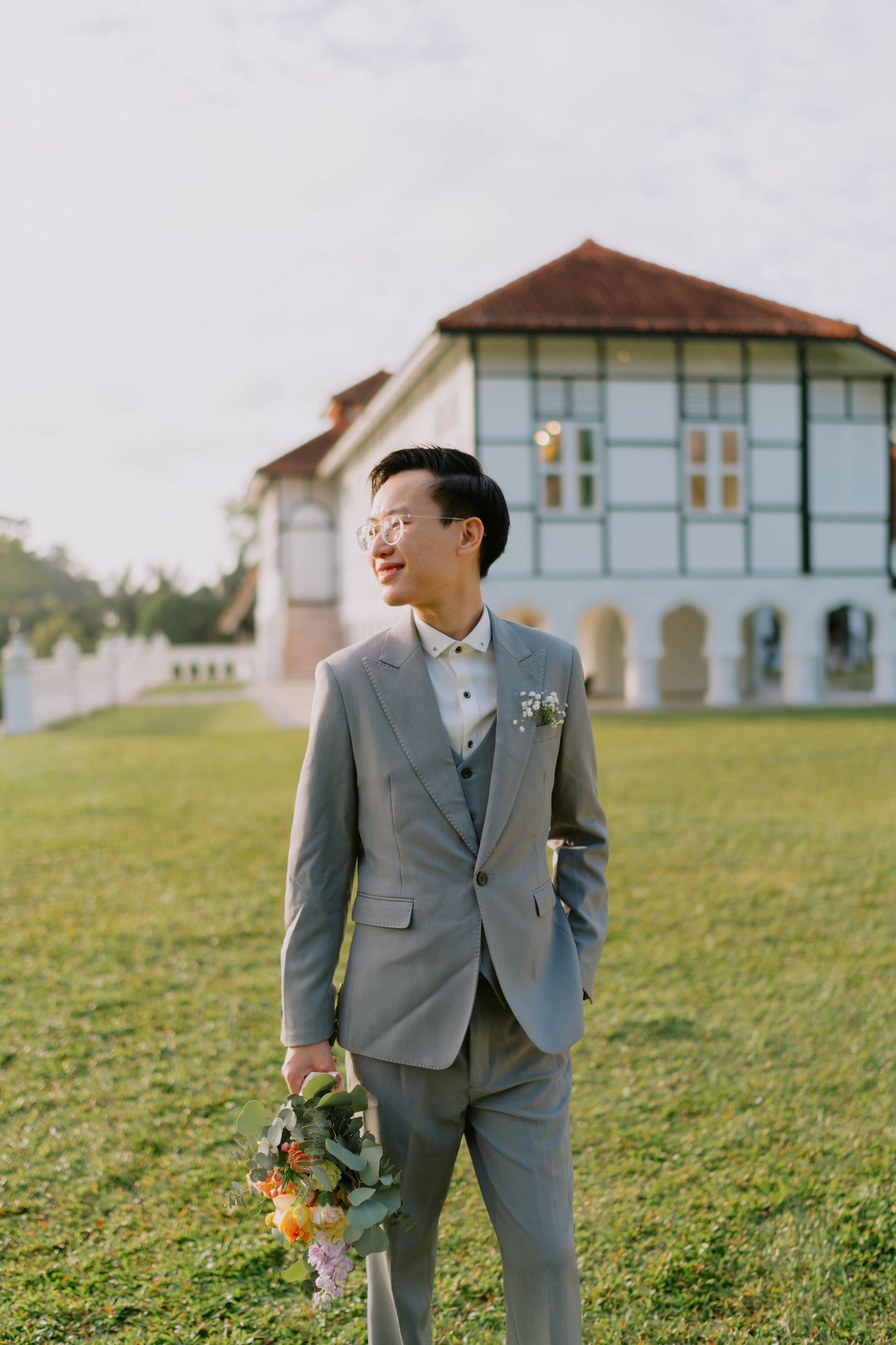 Dapper Asian groom in gray suit with boutonniere holds bouquet while gazing away on green lawn.
