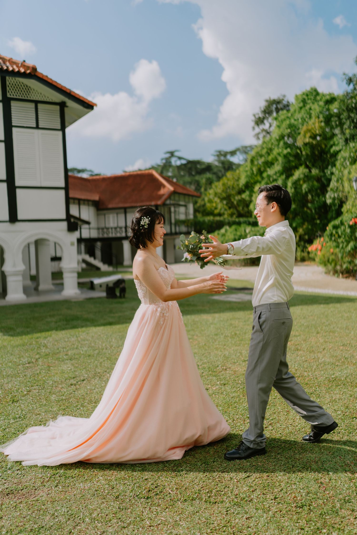 Bride in pink gown and groom playfully reaching for bouquet on lush green lawn outdoors.