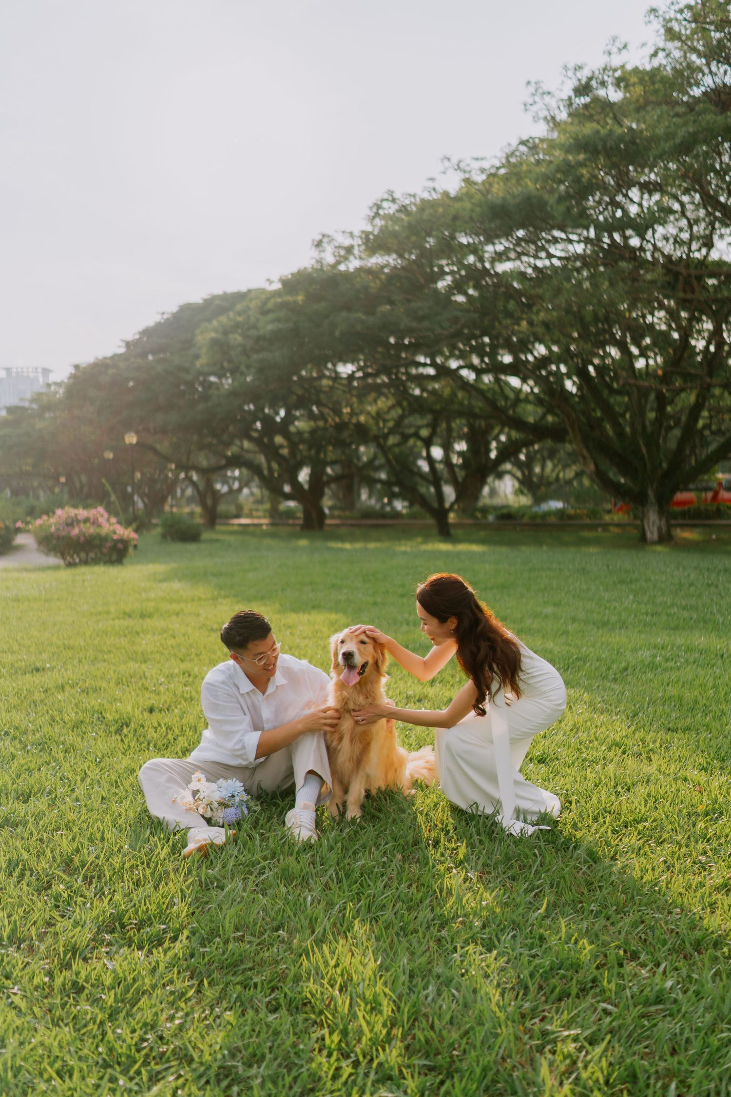 Couple in white outfits sitting on grass petting their golden retriever in a lush green park.
