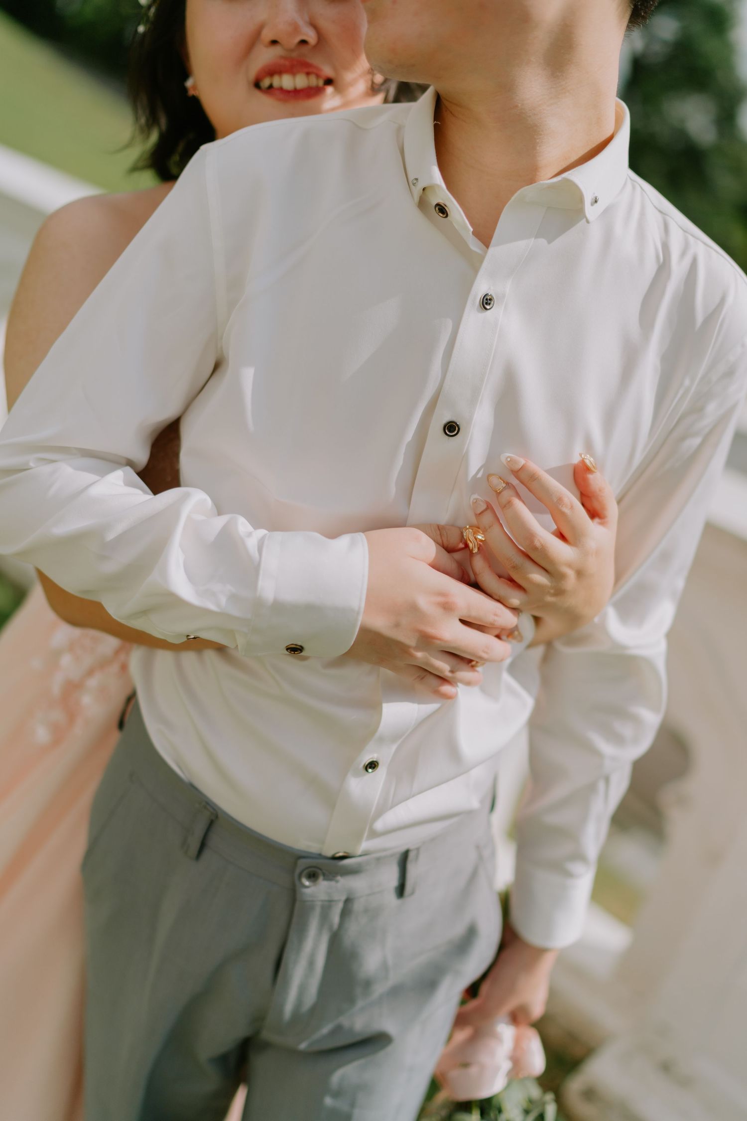 Bride in pink gown embraces groom in white shirt from behind during outdoor wedding photoshoot.
