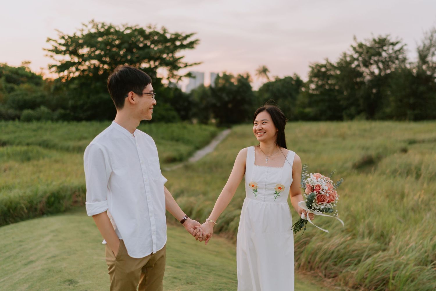 Smiling couple holding hands in a meadow at sunset, woman holding a colorful bouquet.