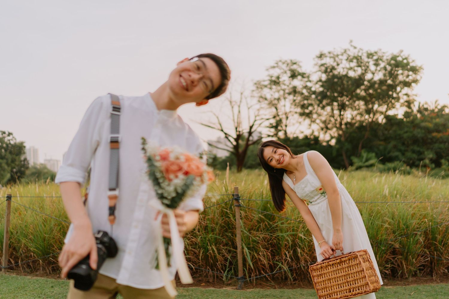 Laughing couple in a green field at dusk, man holding bouquet and camera, woman with picnic basket.