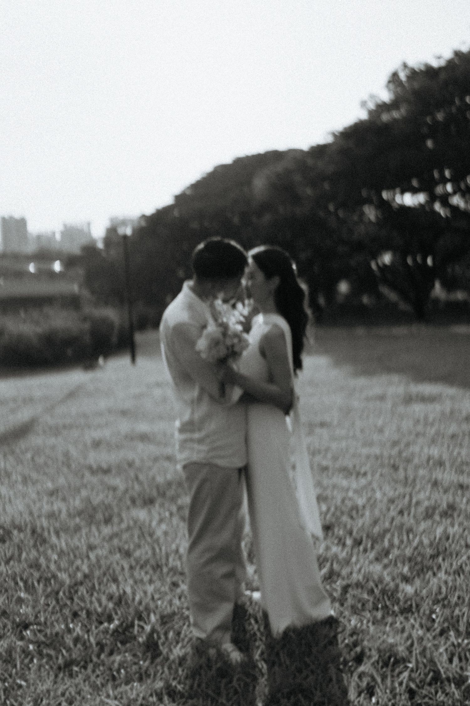 Black and white photo of couple embracing in a park holding a bouquet at dusk.