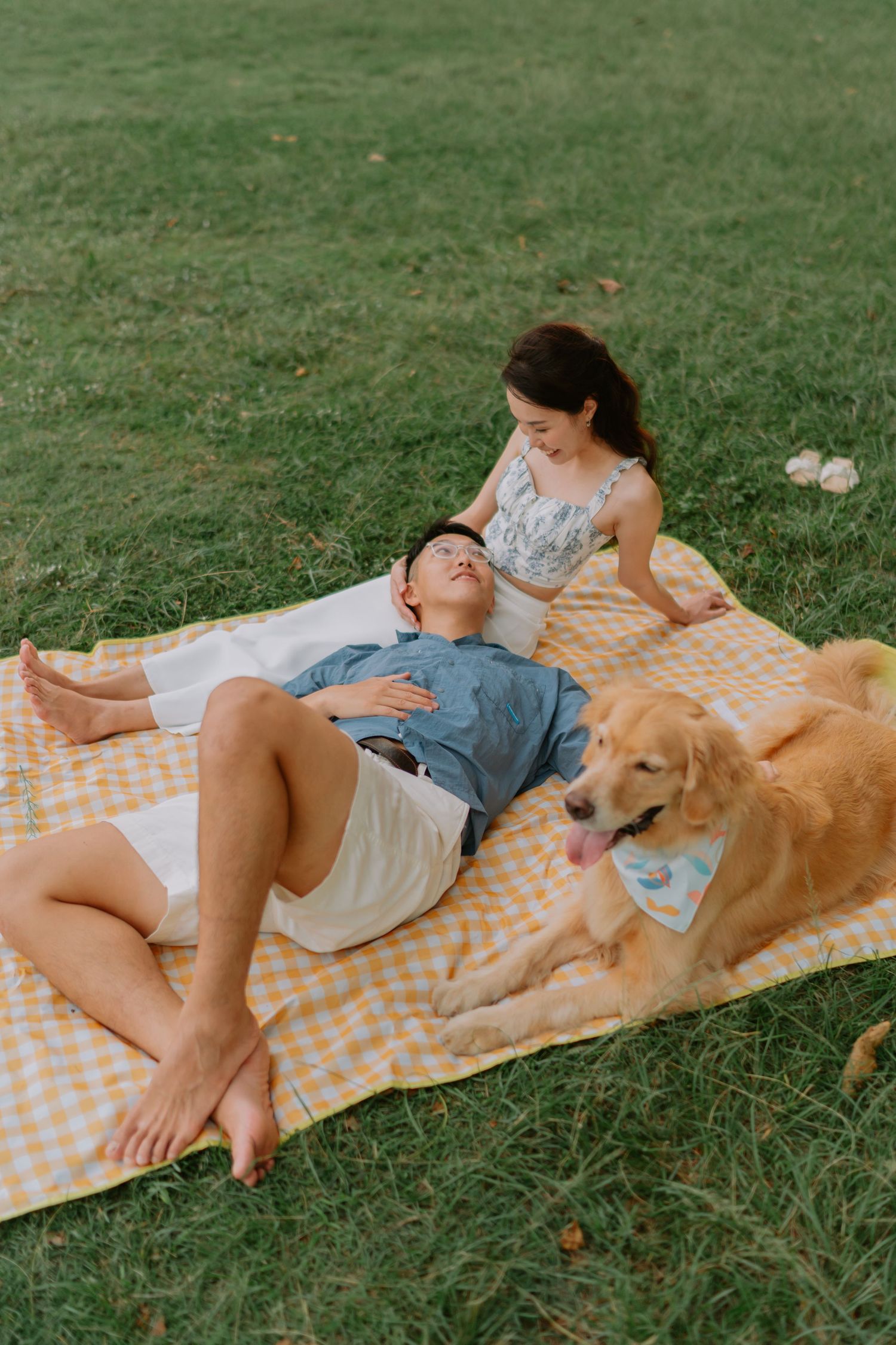 Couple relaxing on yellow checkered picnic blanket with golden retriever on green grass.