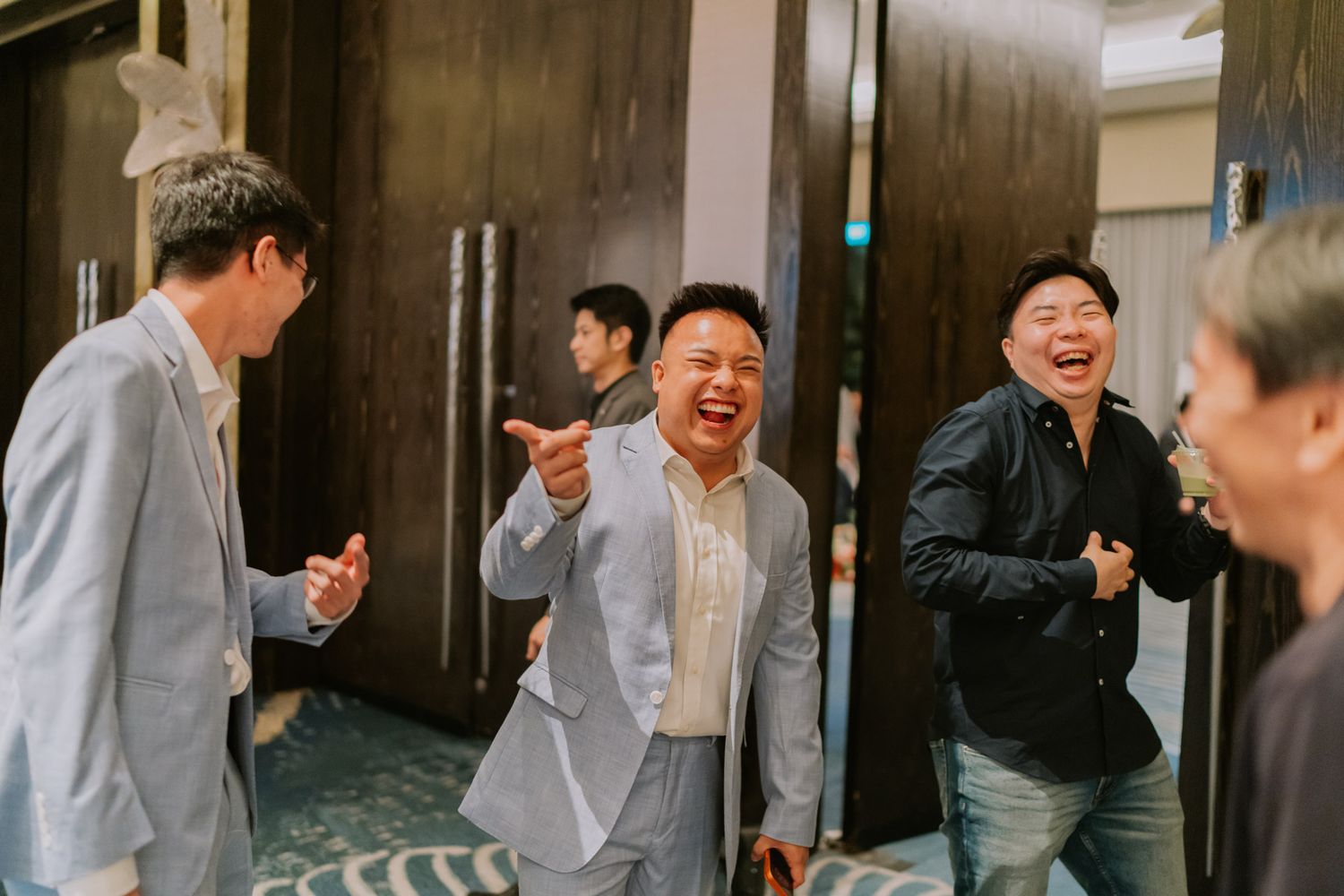 Group of men laughing and celebrating together in a hotel corridor, wearing smart casual attire at a social event.