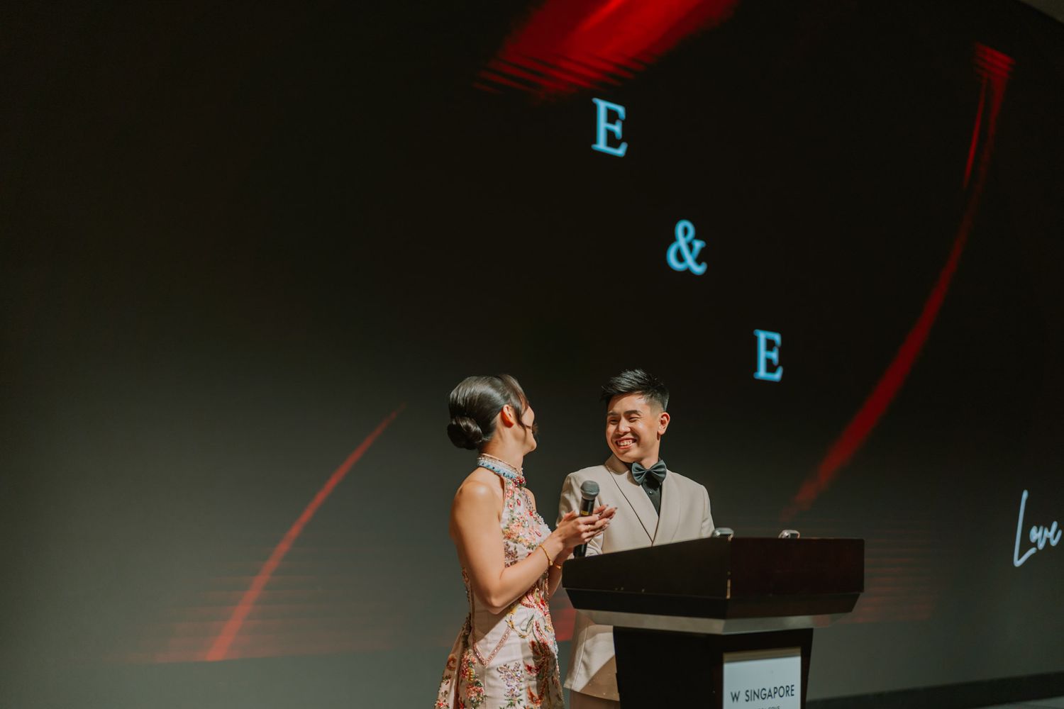 Couple sharing a romantic moment at a wedding reception podium with E&E monogram projected on screen behind them.