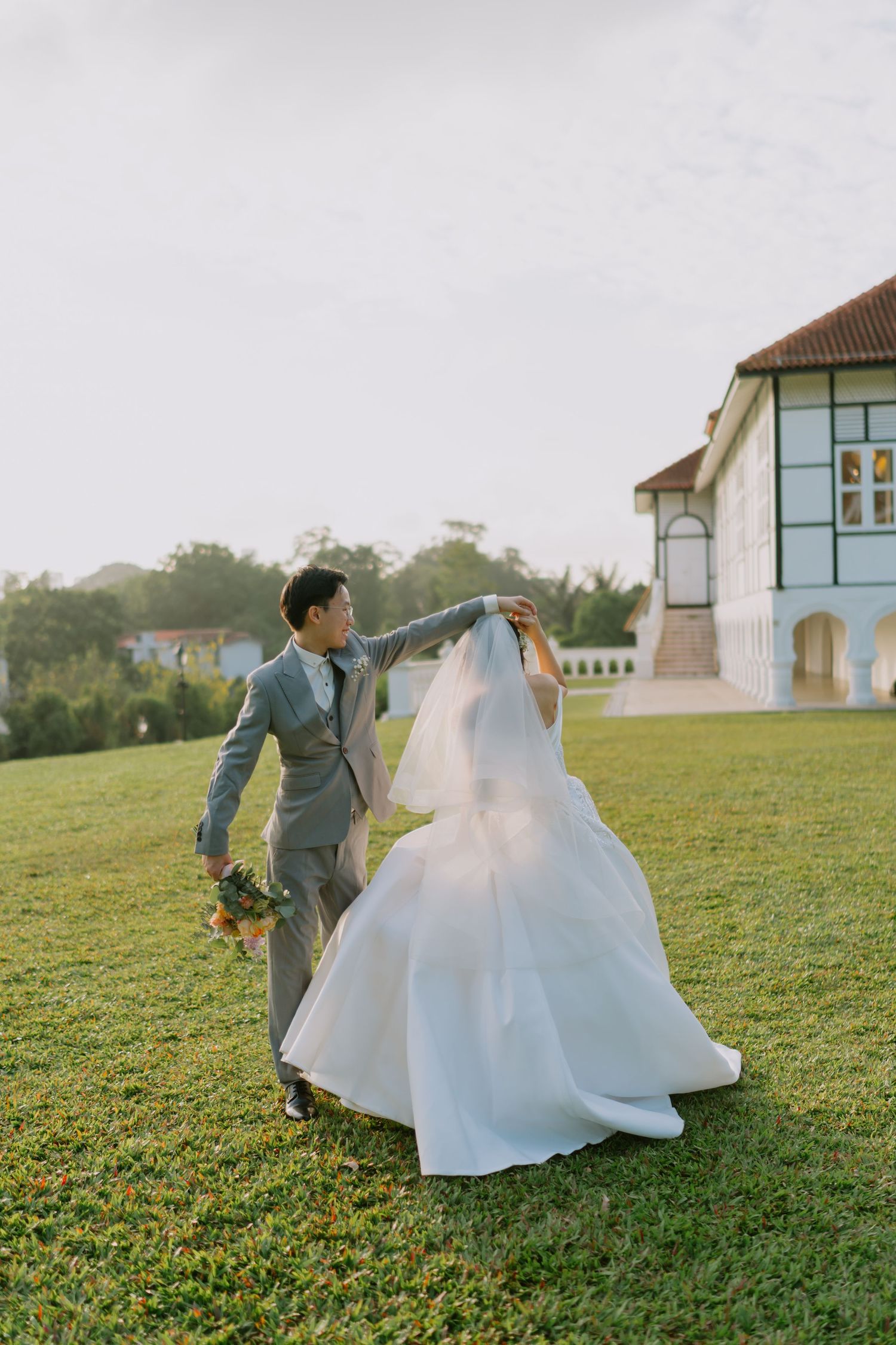 Groom in gray suit lifts bride's veil on green lawn beside white colonial-style building.