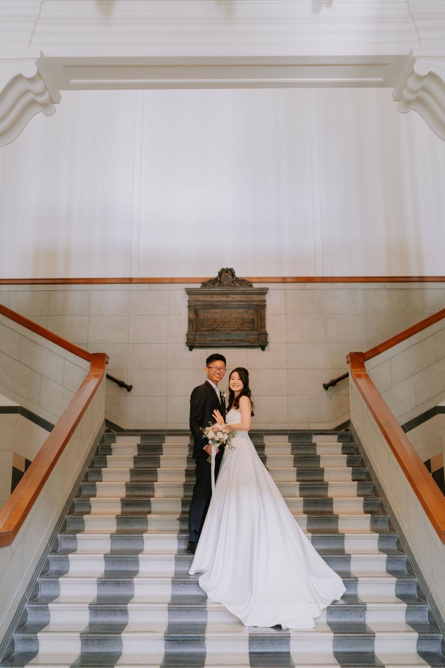 Wedding couple posing on grand staircase, bride in flowing white gown holding bouquet.