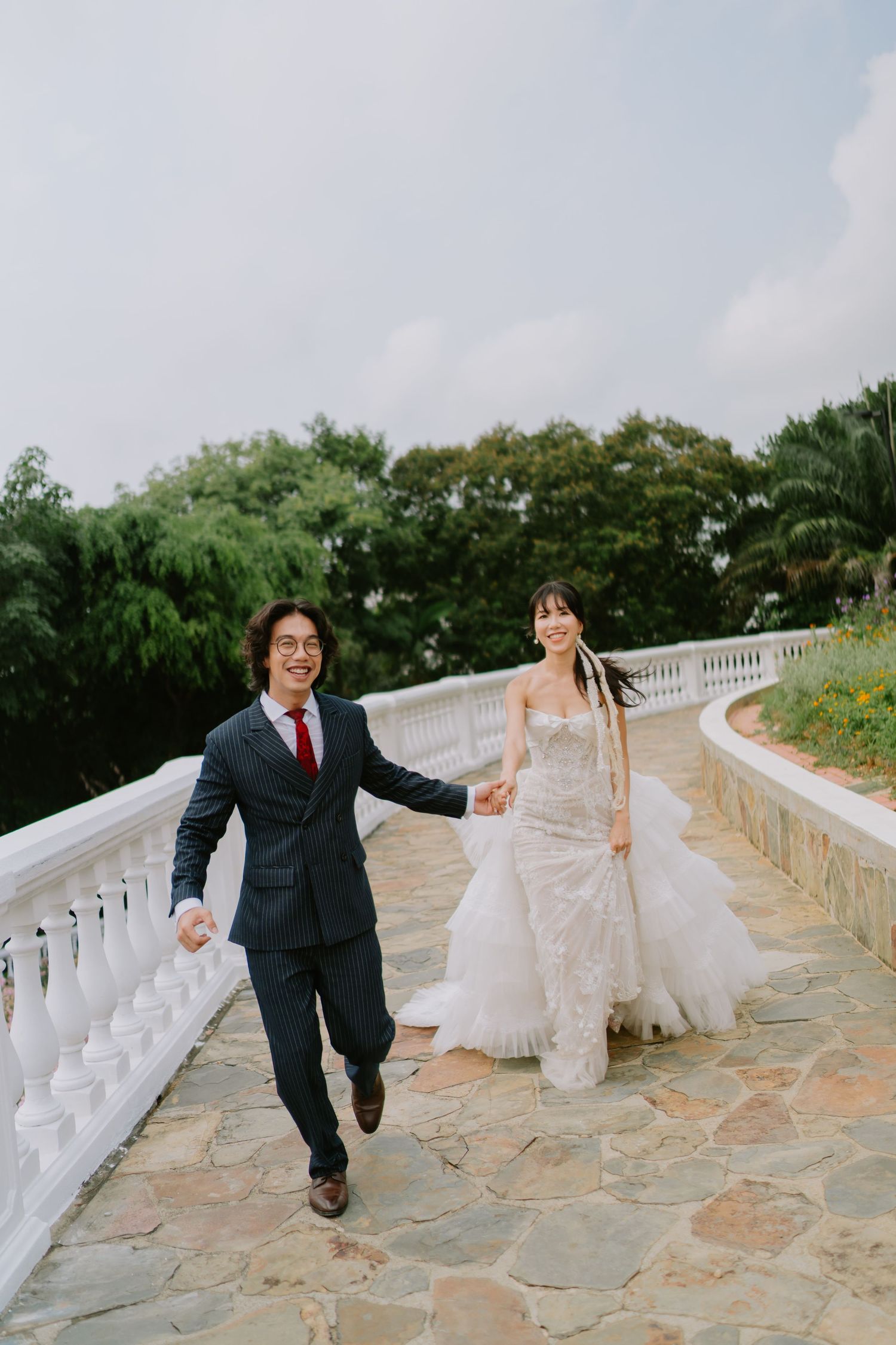 Happy bride and groom holding hands and walking along a white balustrade terrace with green trees behind them.