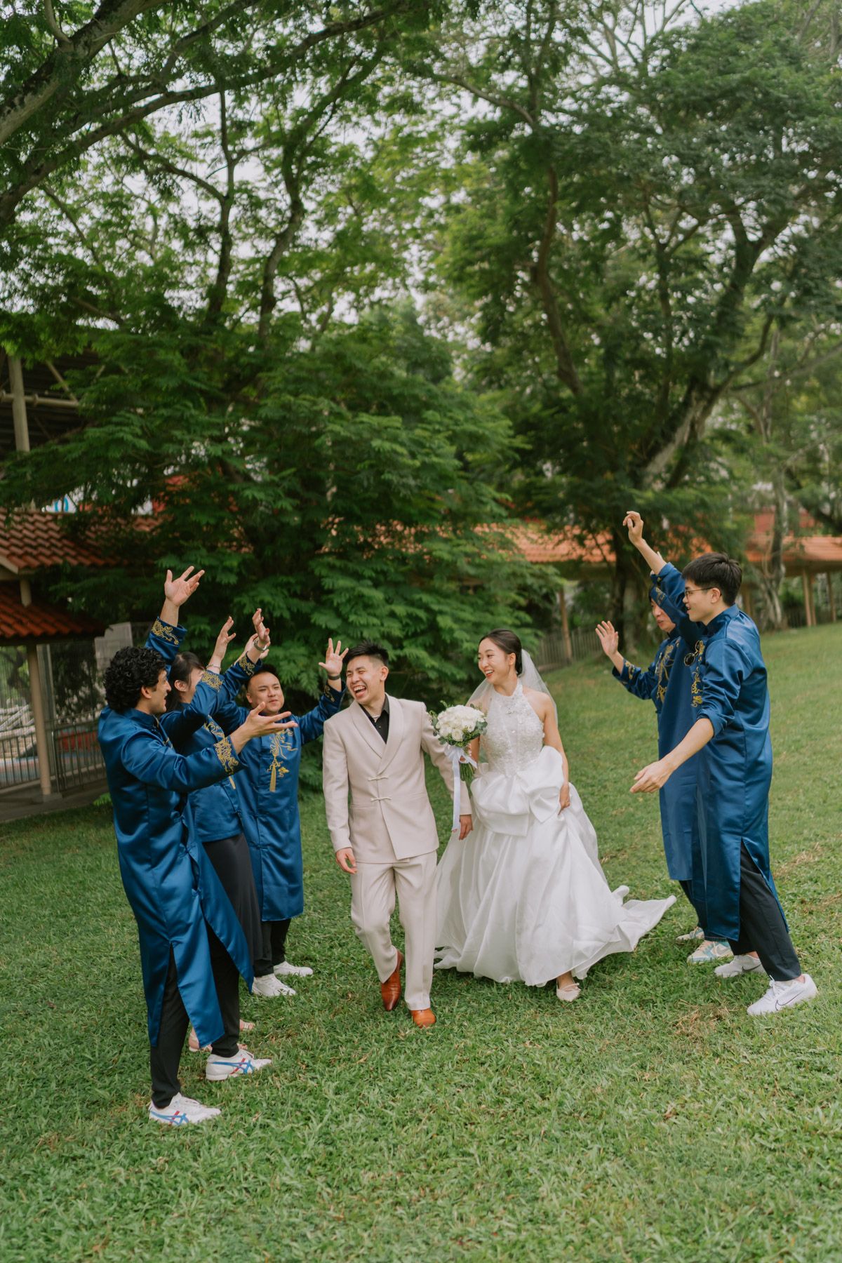 Bride and groom walk through cheering groomsmen in blue suits on green lawn with lush trees backdrop.