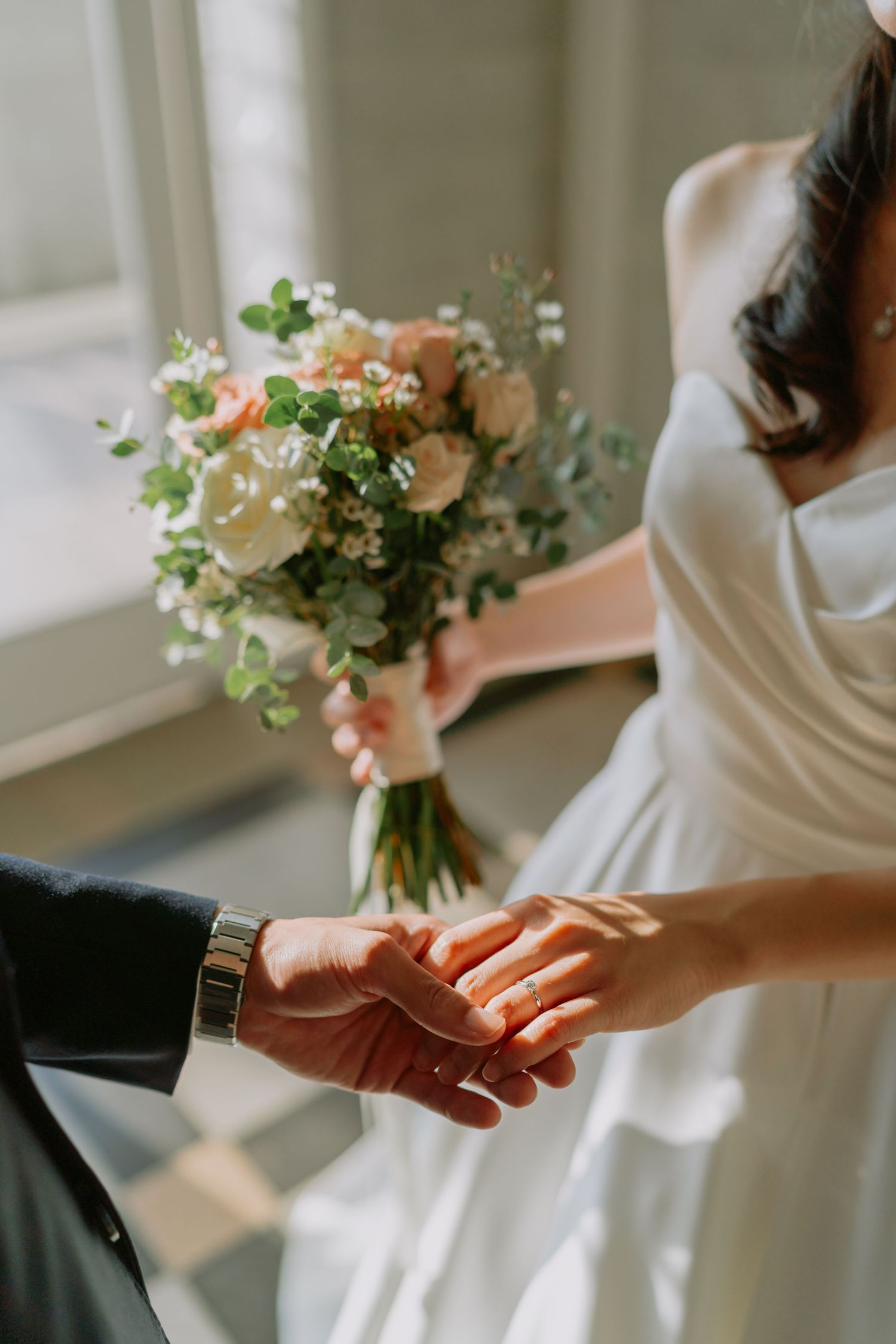 Close-up of bride and groom holding hands, bride holding white and pink floral wedding bouquet.