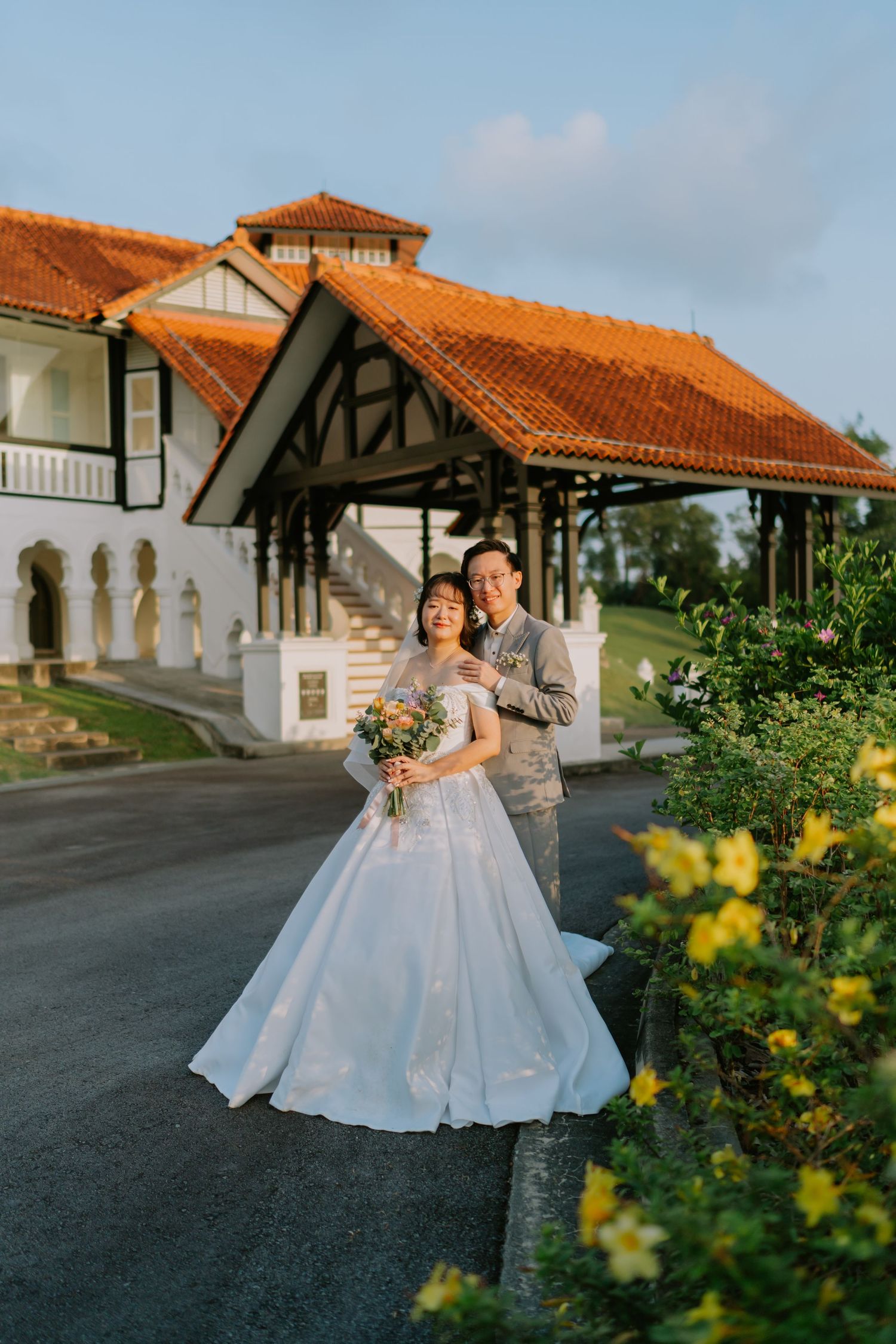 Asian wedding couple poses in front of colonial building with red tile roof surrounded by lush greenery.