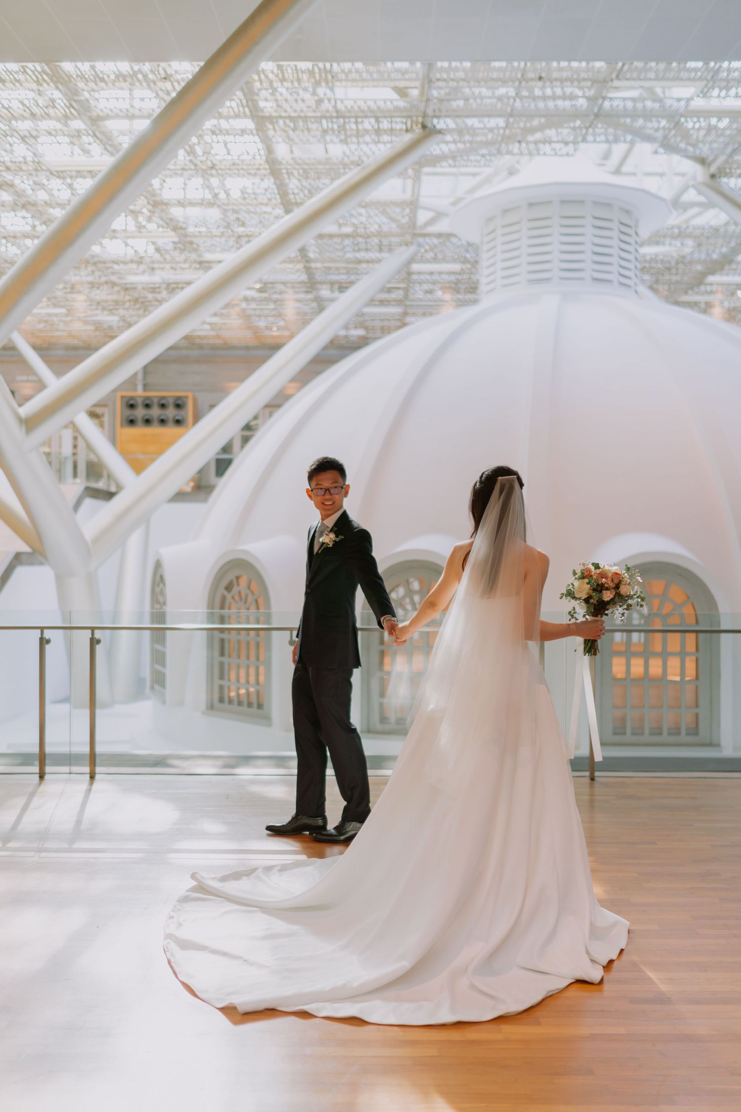 Full-length shot of Asian wedding couple holding hands in elegant white domed venue, bride's long veil trailing.