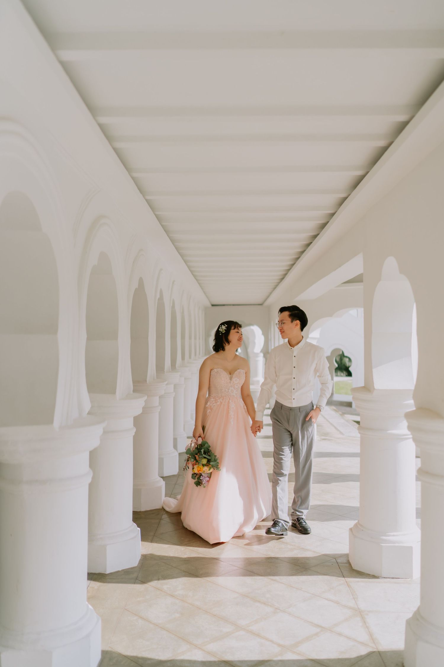 Bride in pink gown and groom walking hand in hand through a sunlit white arched corridor.