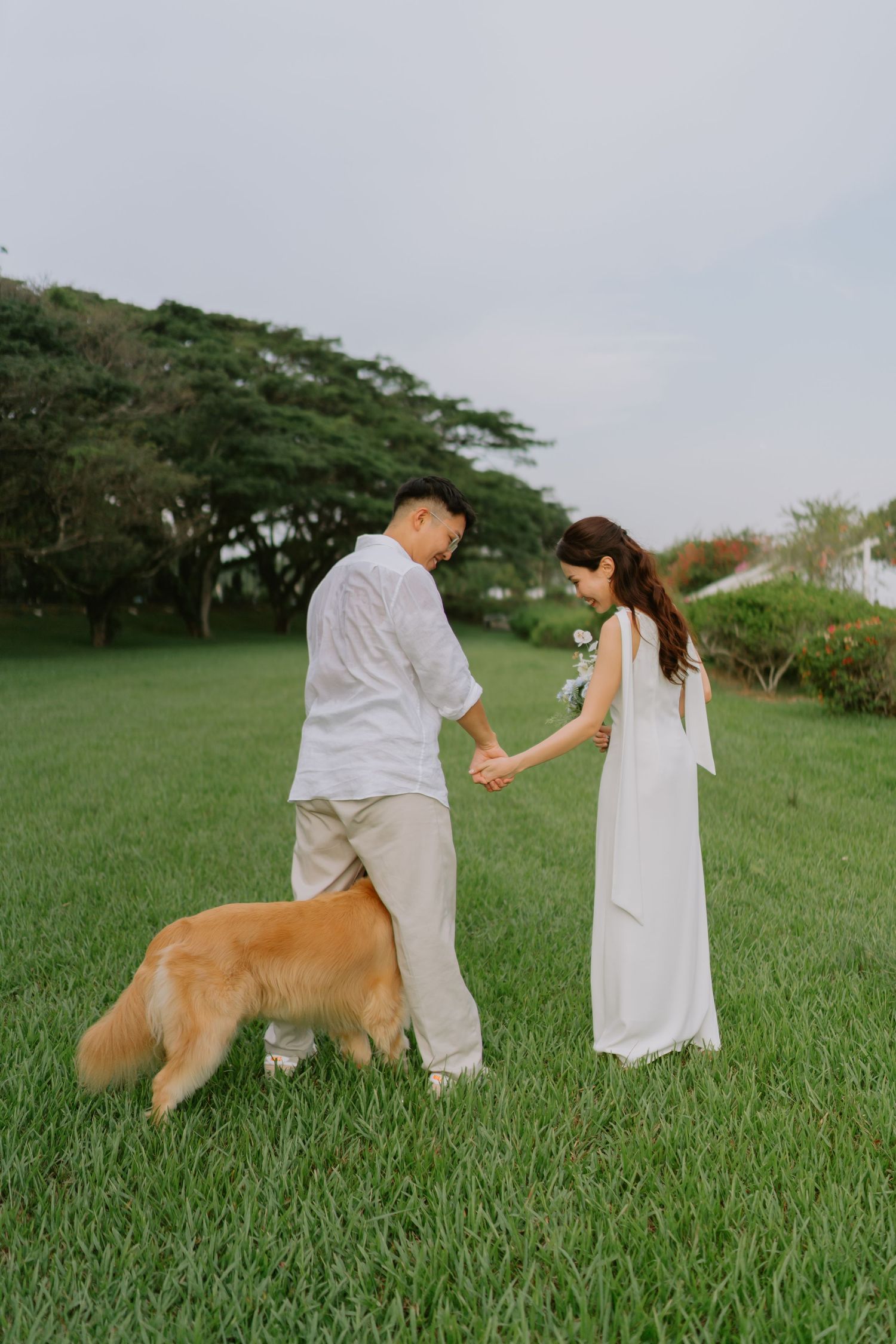 Wedding couple in white attire hold hands and look down at golden retriever on green lawn.