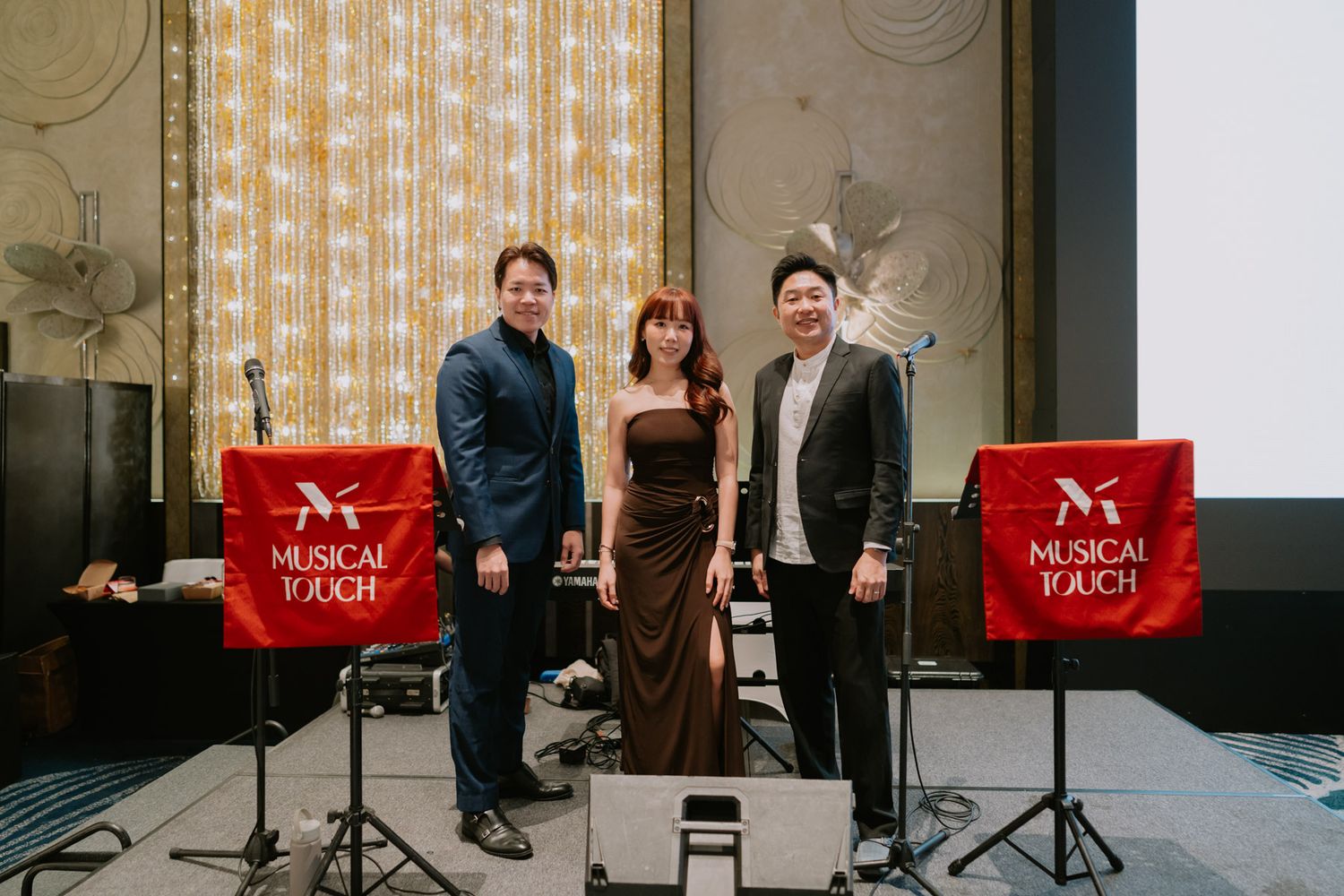 Three performers pose between two Musical Touch branded red stands at an elegant indoor event venue.
