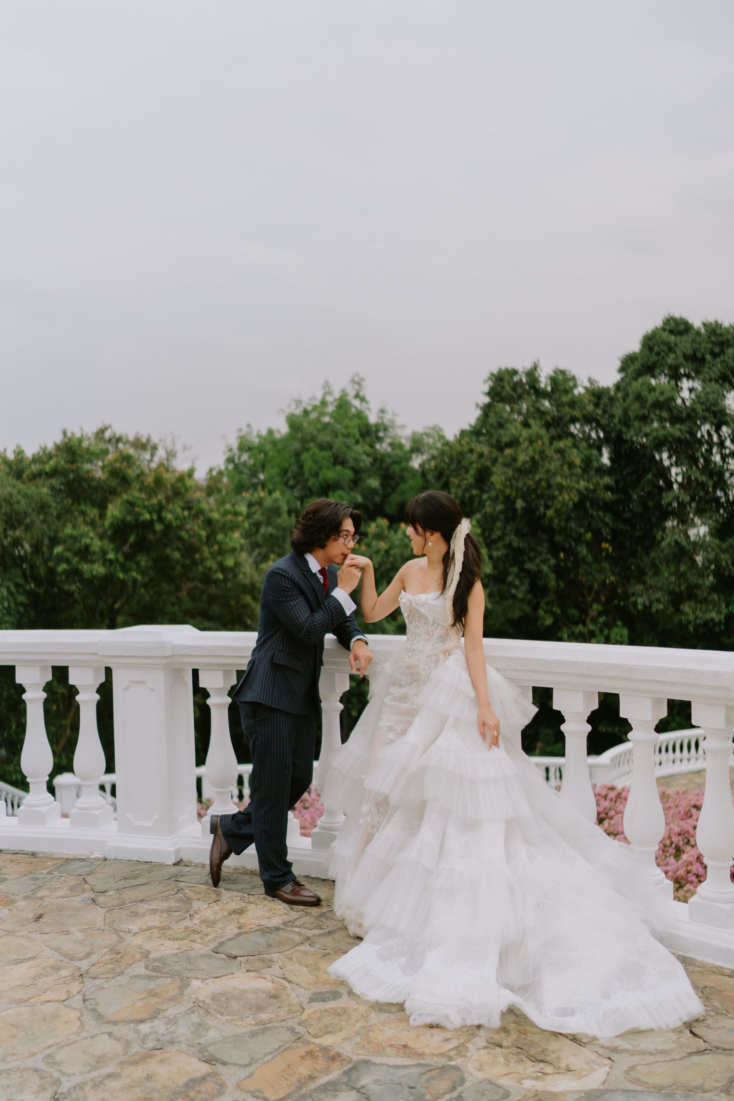 Groom kissing bride's hand on a white balustrade terrace surrounded by lush green trees and pink flowers.