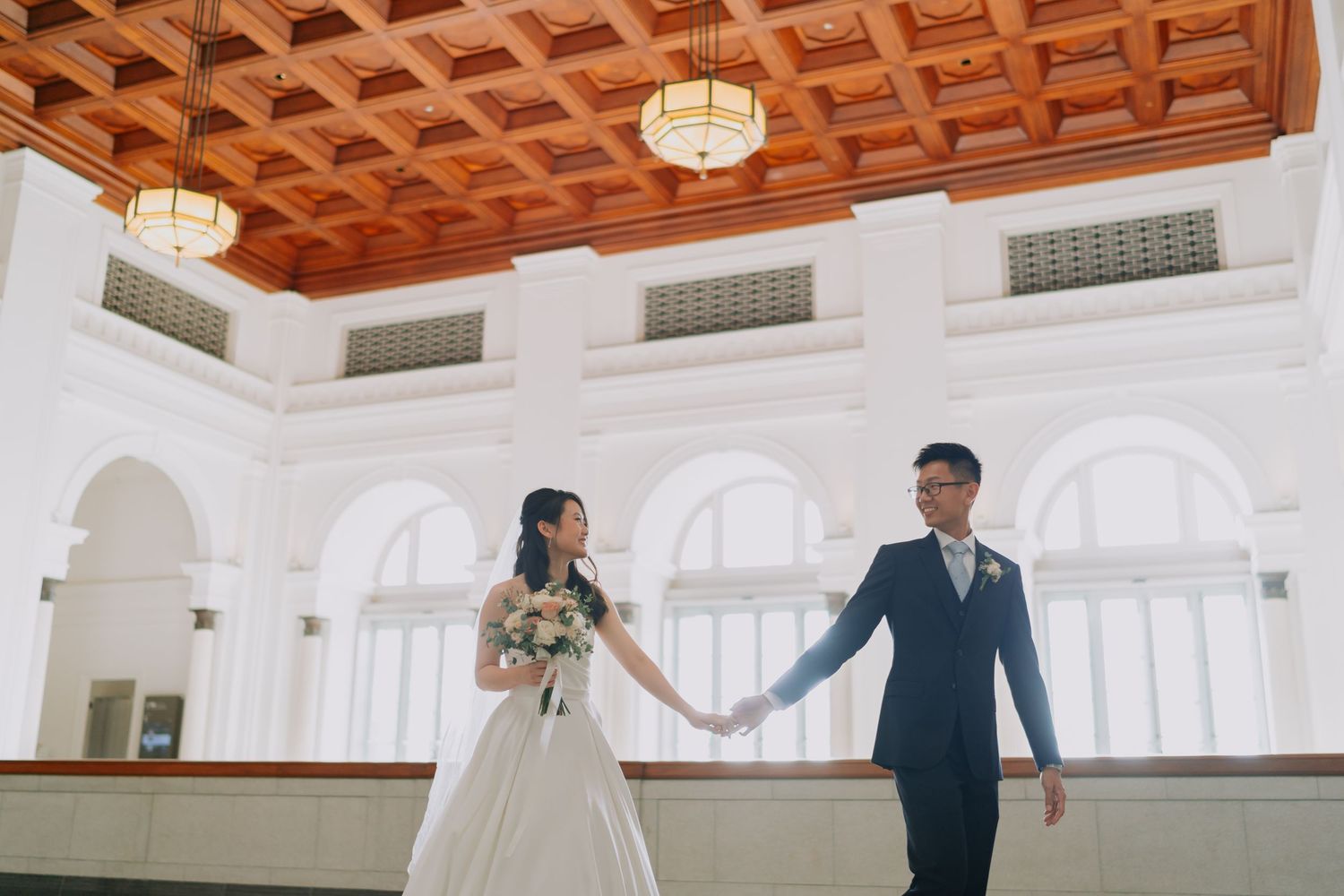 Bride and groom holding hands walking in grand white hall with ornate wooden ceiling.