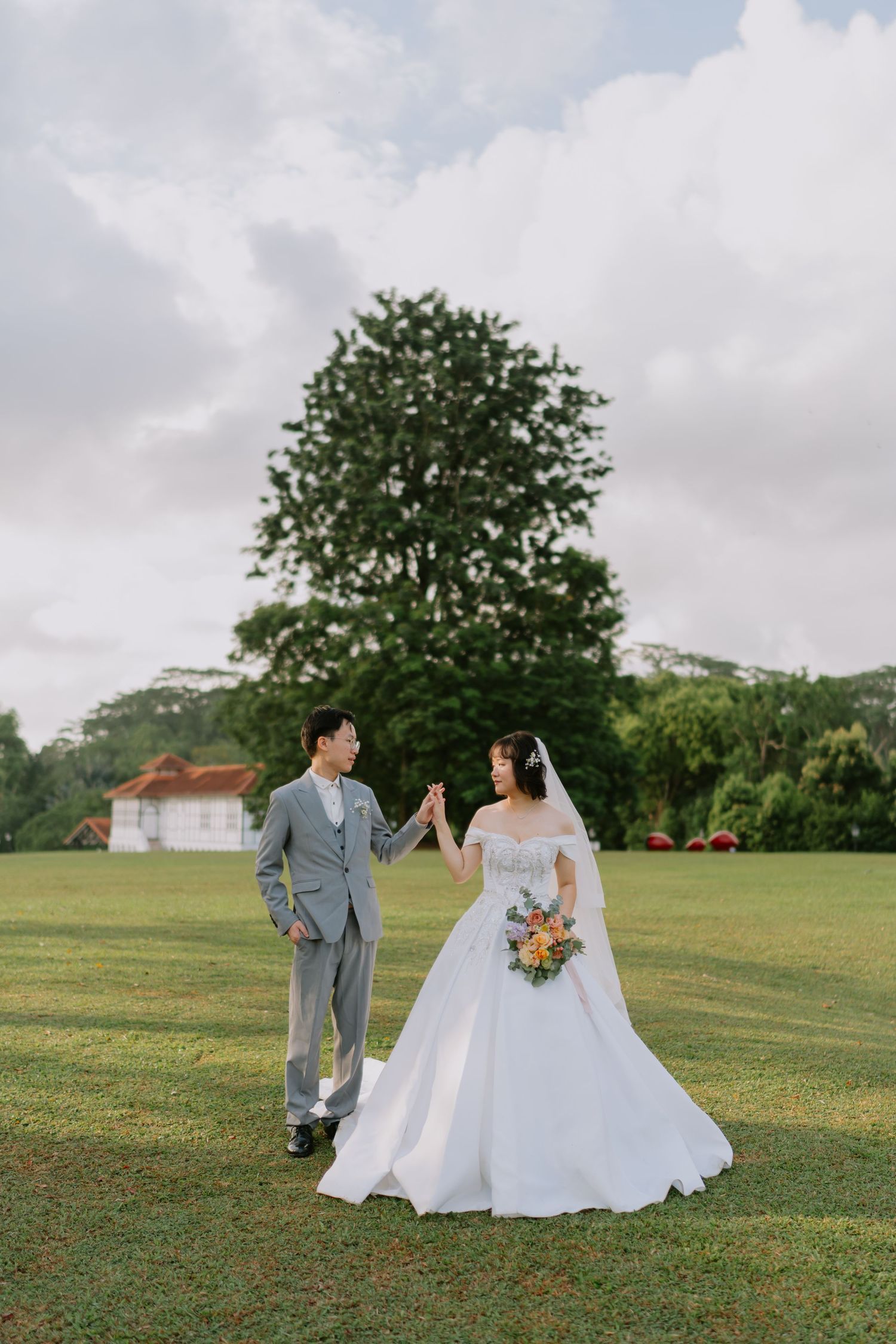 Bride and groom hold hands while walking across a green field with a large tree in the background.