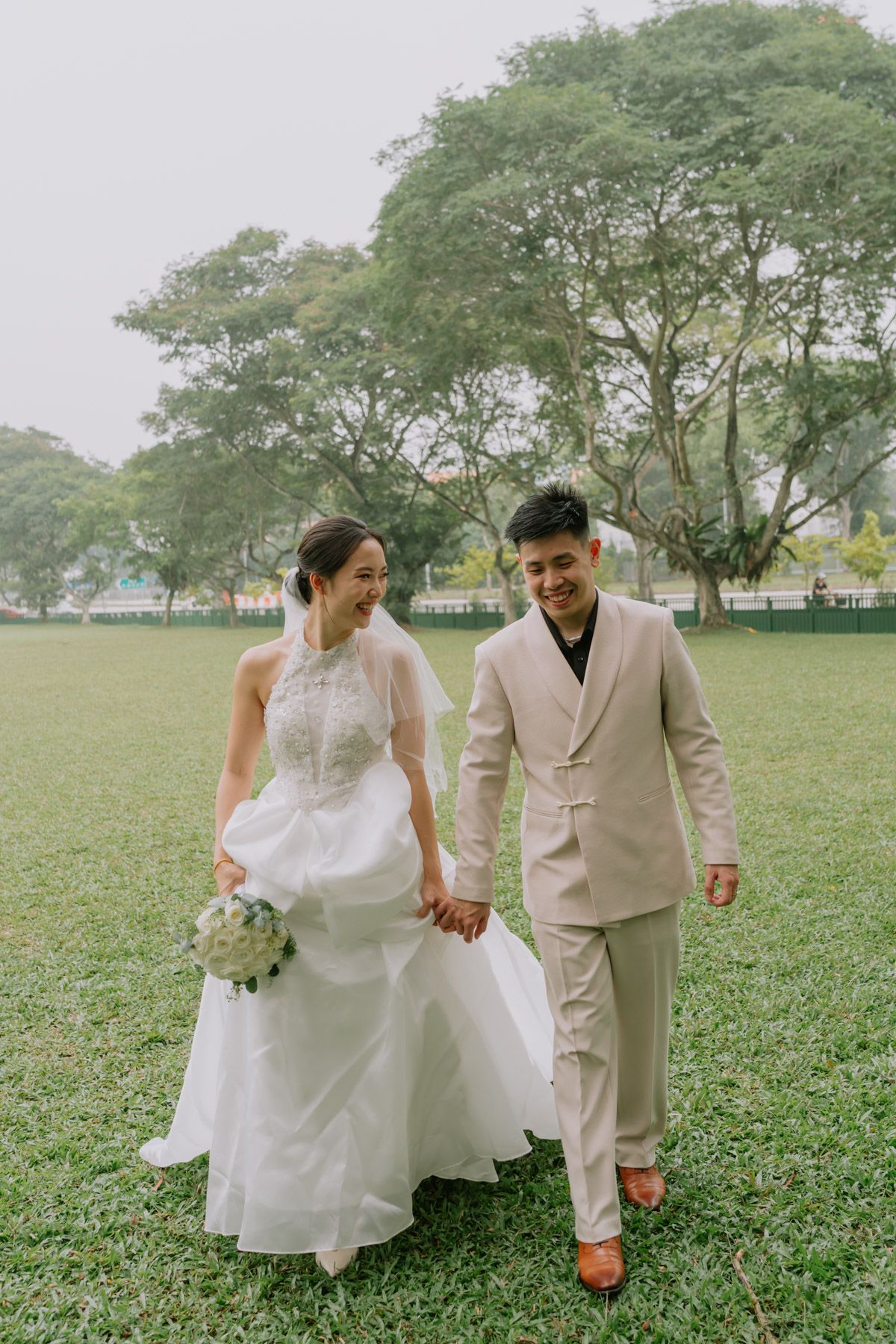 A couple in wedding attire walks hand-in-hand on a green lawn, bride in white gown holding bouquet, groom in beige suit.