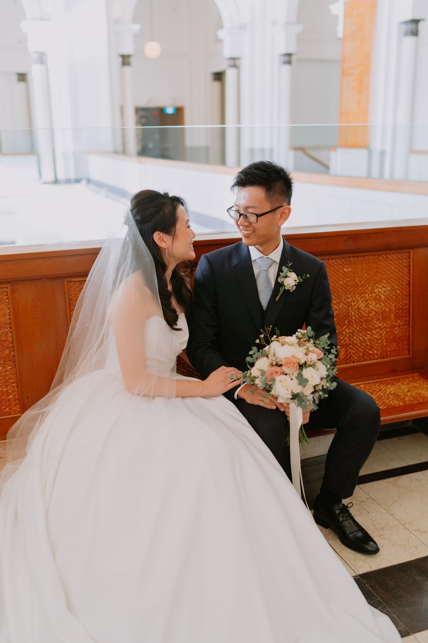 Asian bride and groom sitting on church pew, smiling at each other, bride holding pink and white wedding bouquet.