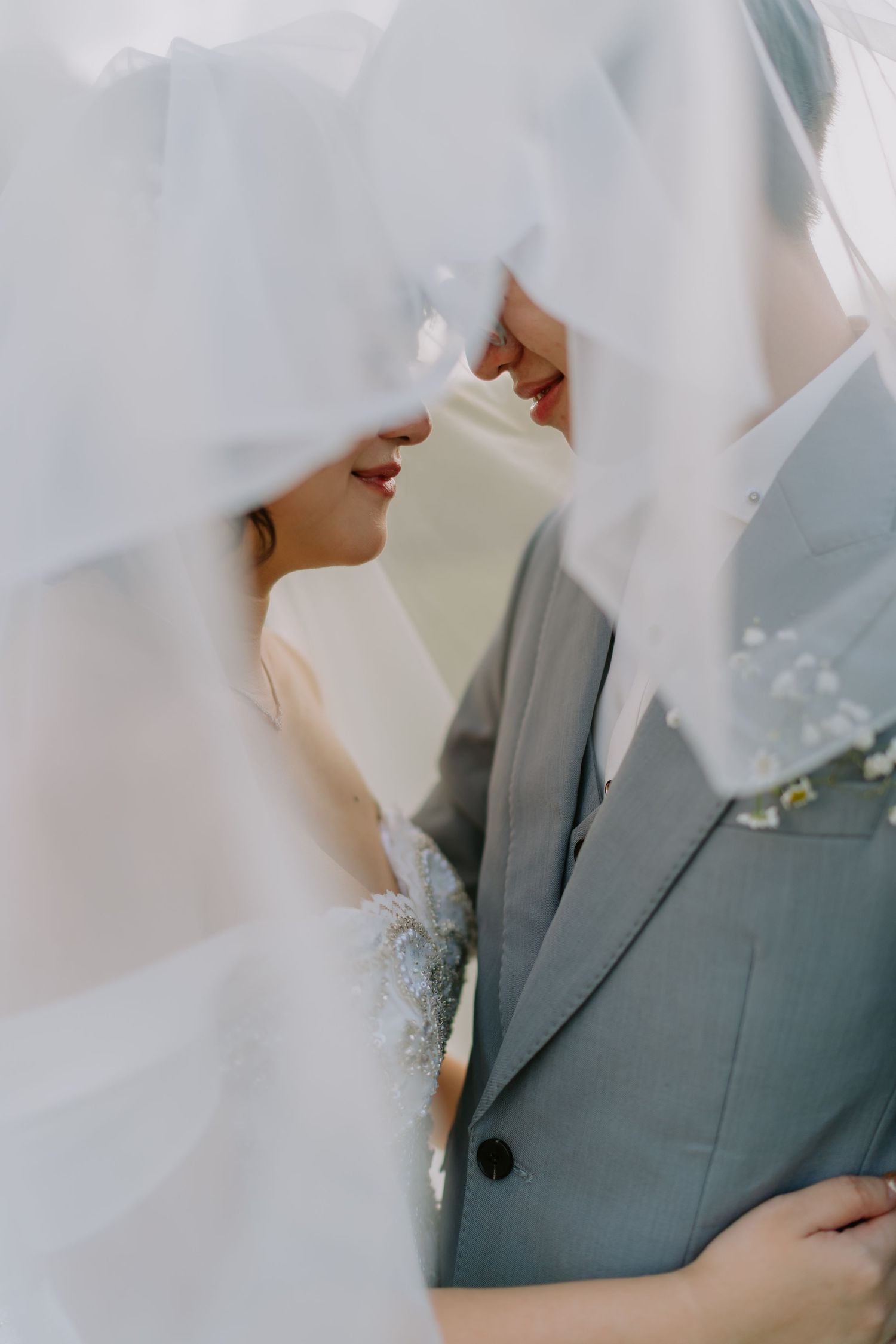 Close-up of bride and groom nearly kissing beneath a flowing white wedding veil in soft light.