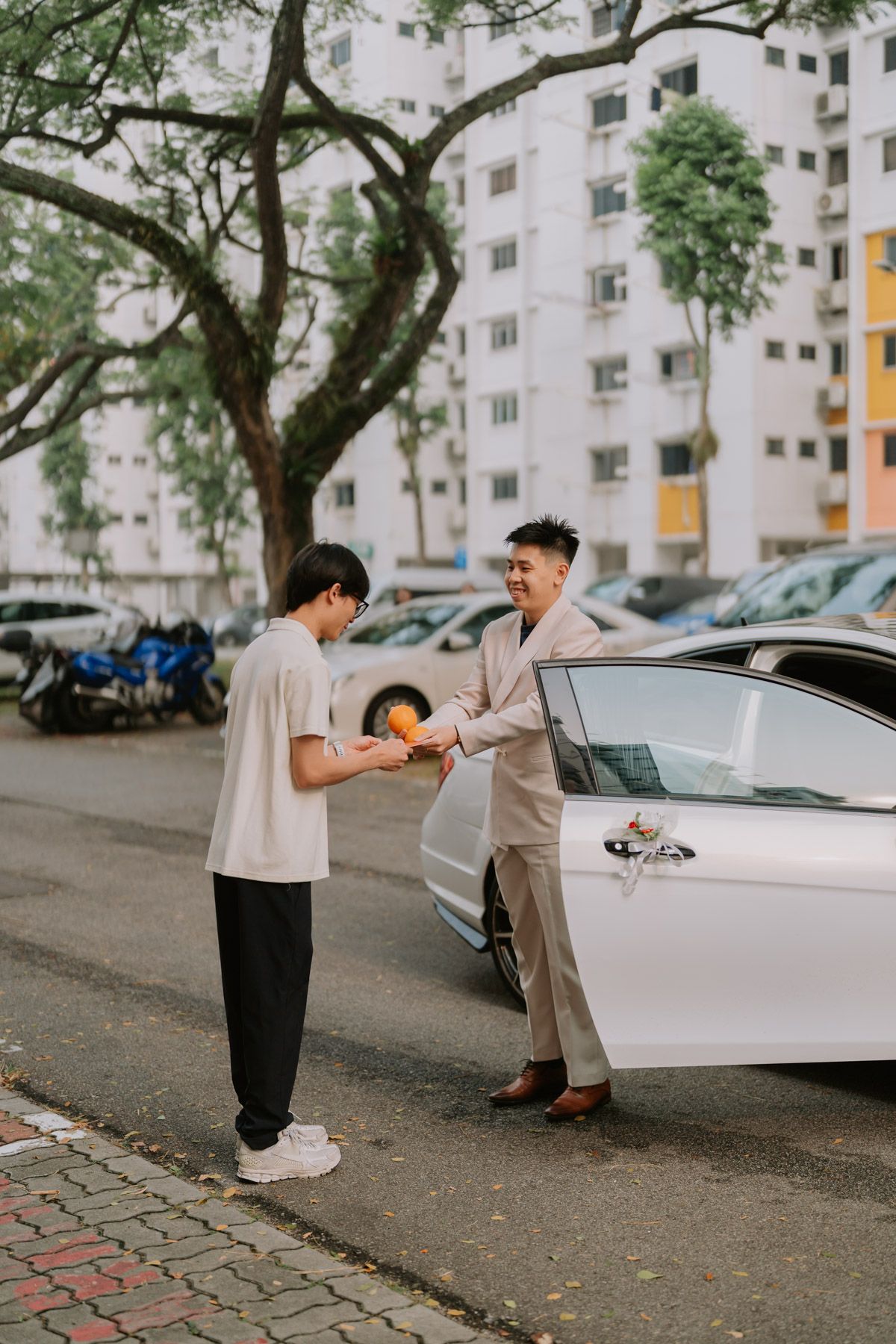 Two Asian men exchange an orange fruit beside a white car in a Singapore HDB estate parking lot.