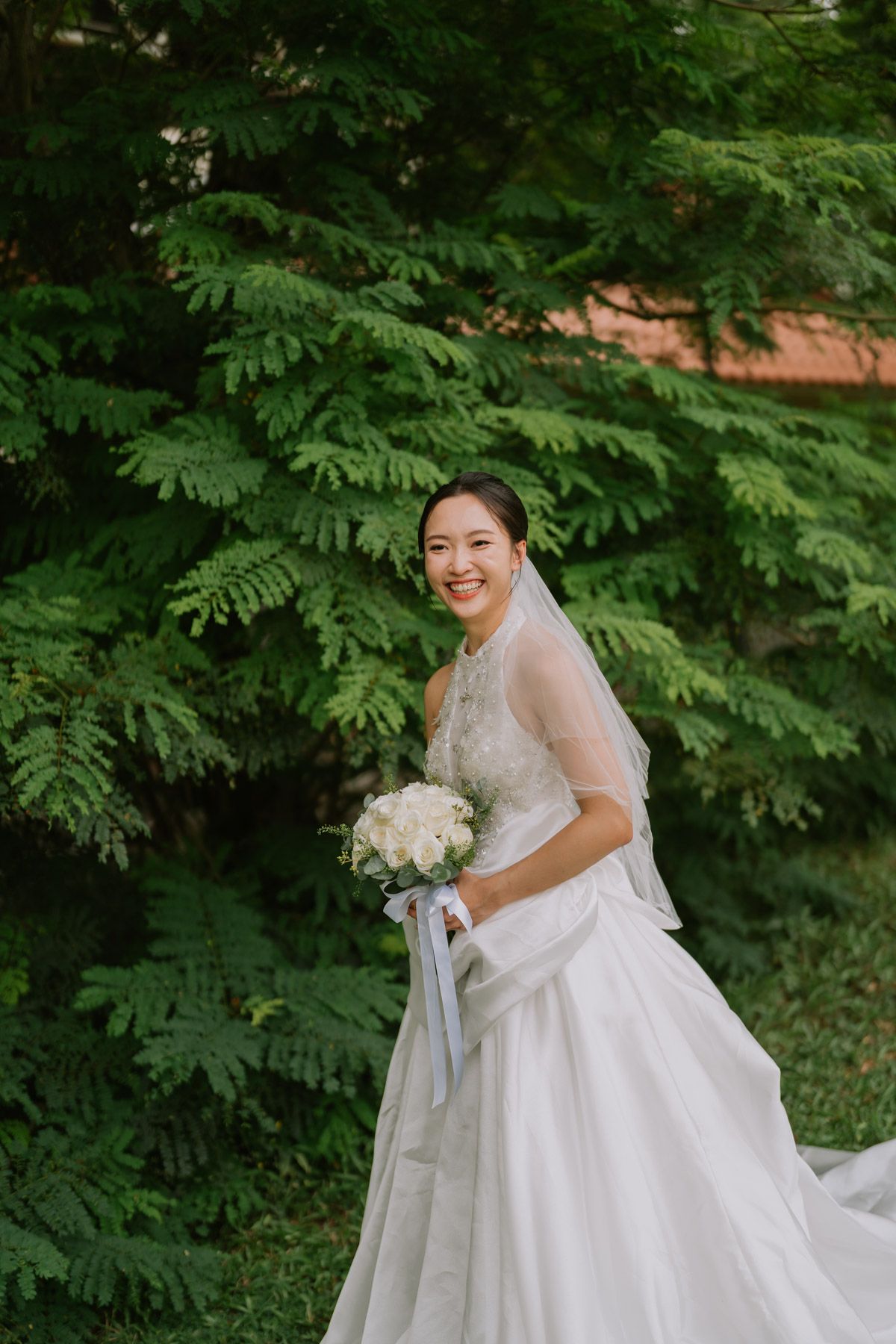 Smiling bride in white gown and veil holds white bouquet outdoors with lush green trees in background.