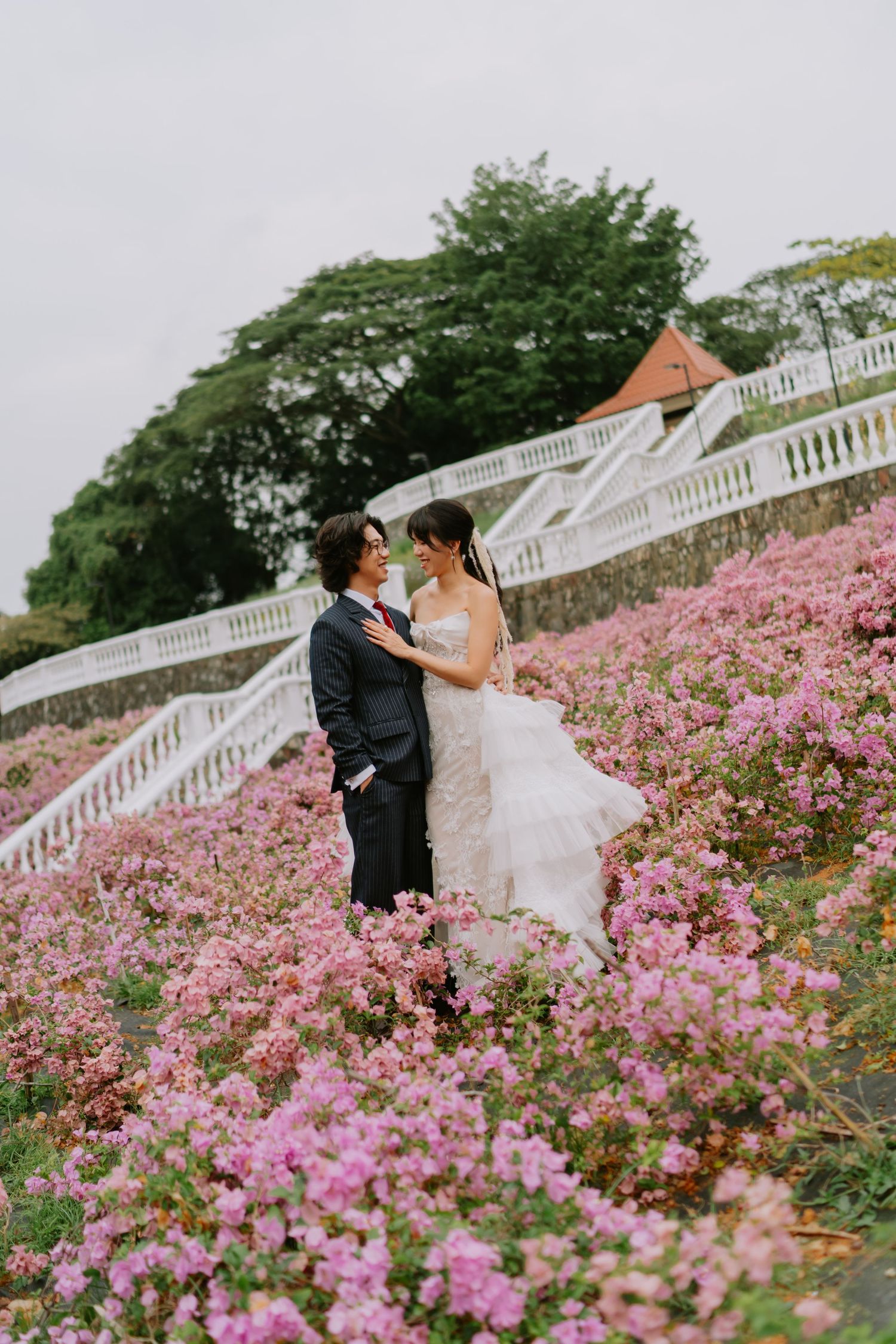 Bride and groom embracing romantically in a field of pink flowers with a white staircase in the background.