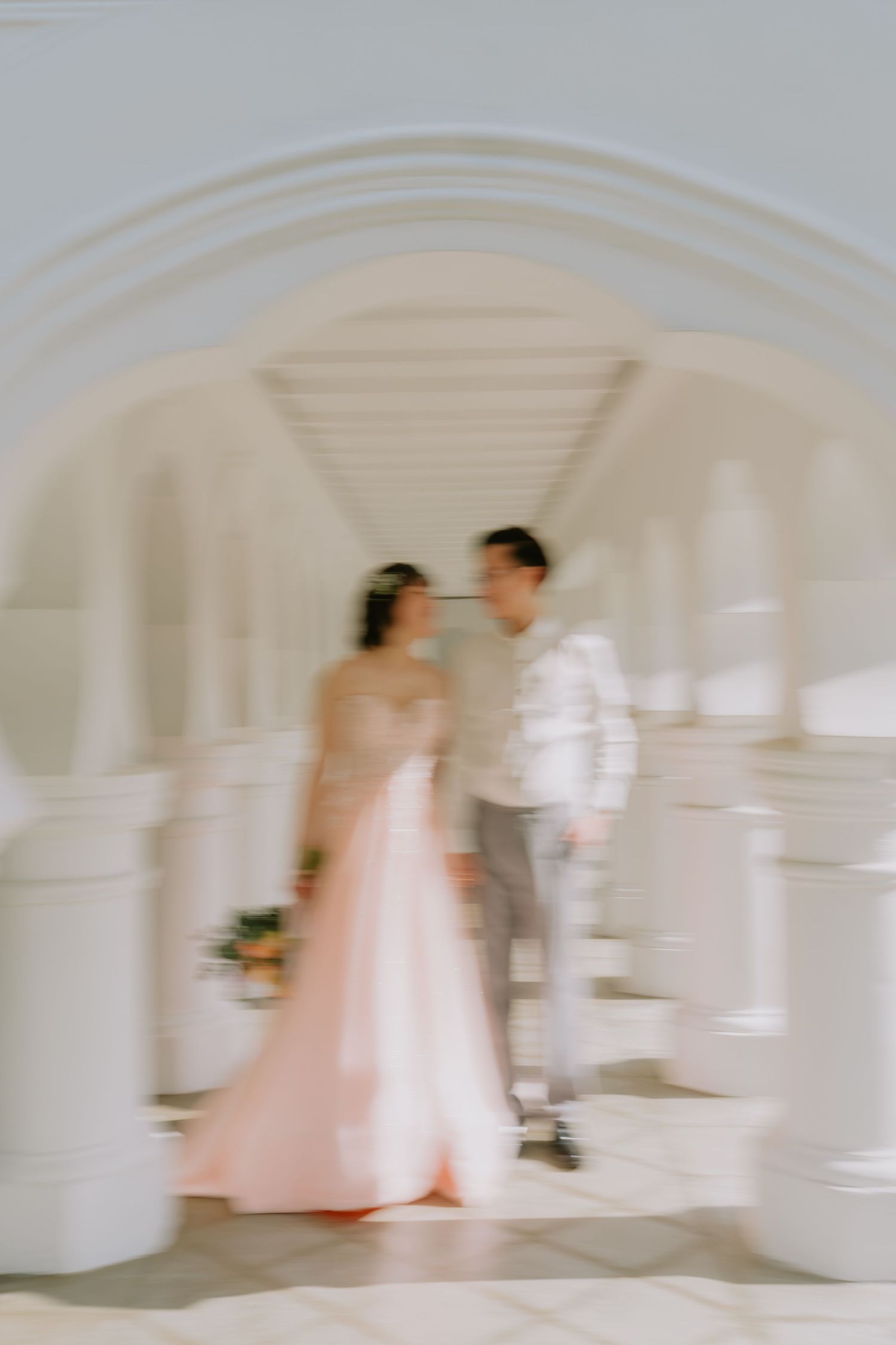 Soft blurred artistic shot of bride in pink gown and groom standing under white arched corridor.