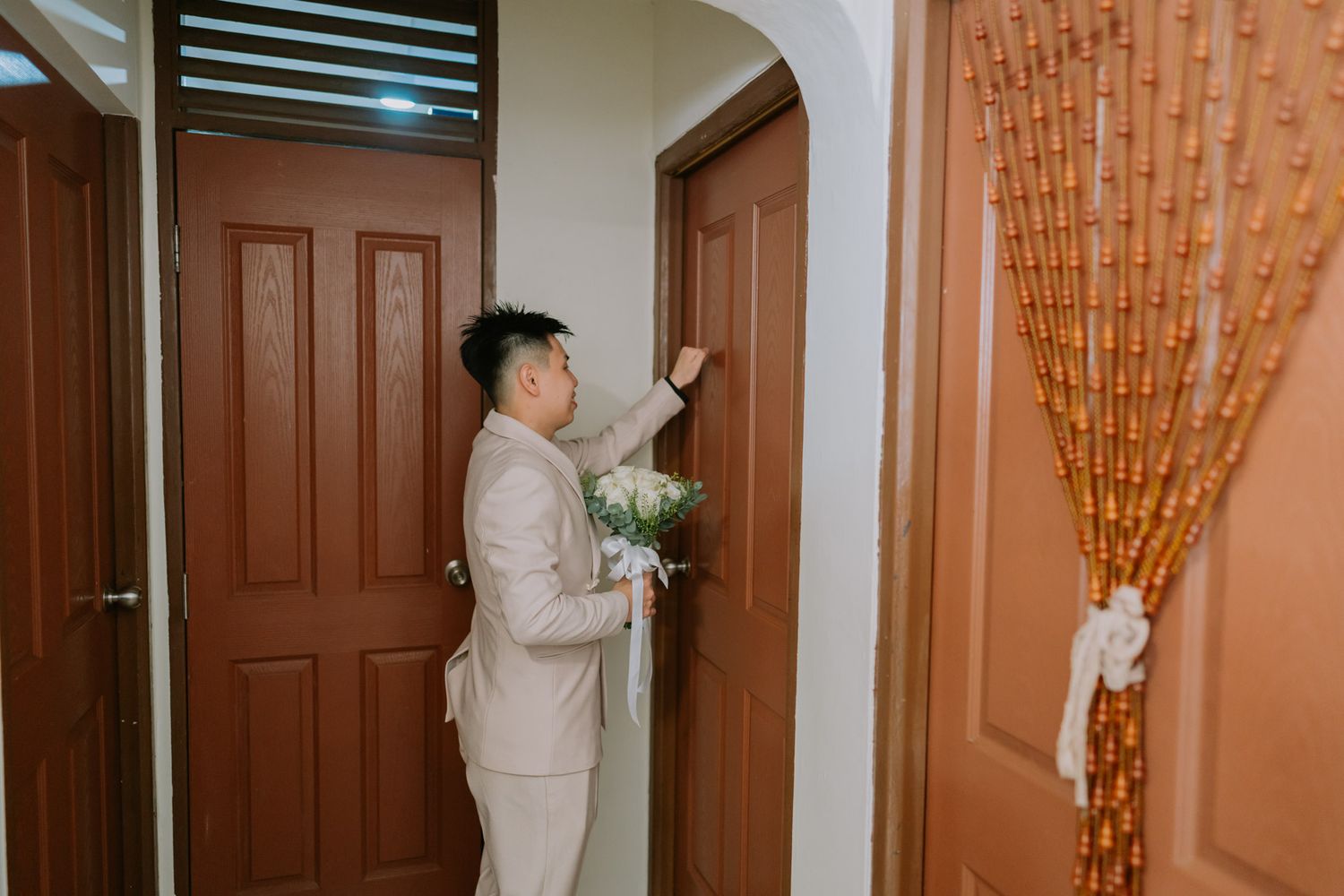 A groom in a beige suit holding a bouquet knocks on a wooden door, with a decorative bamboo curtain nearby.