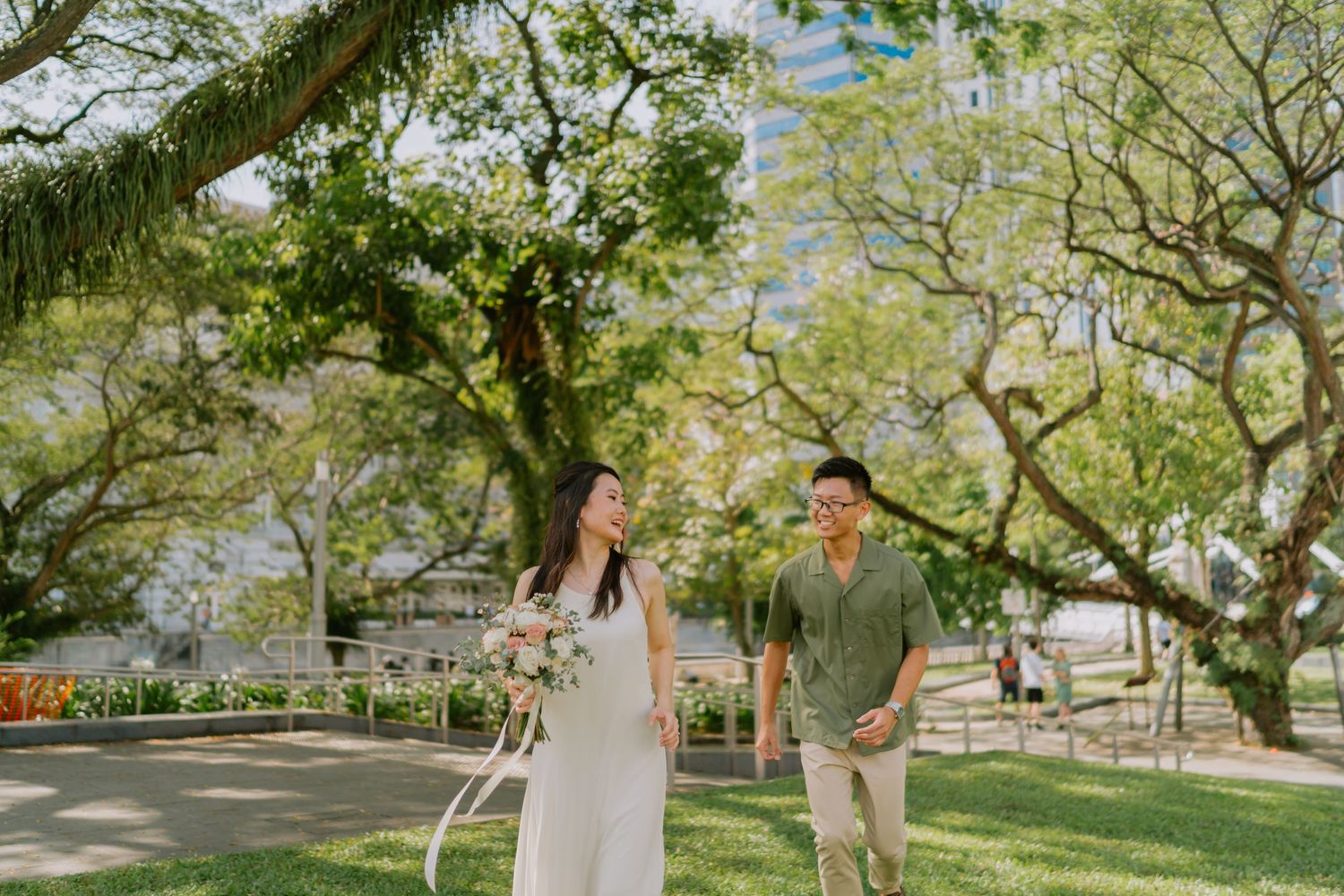 Couple walking through a sunlit park with lush green trees and bouquet.
