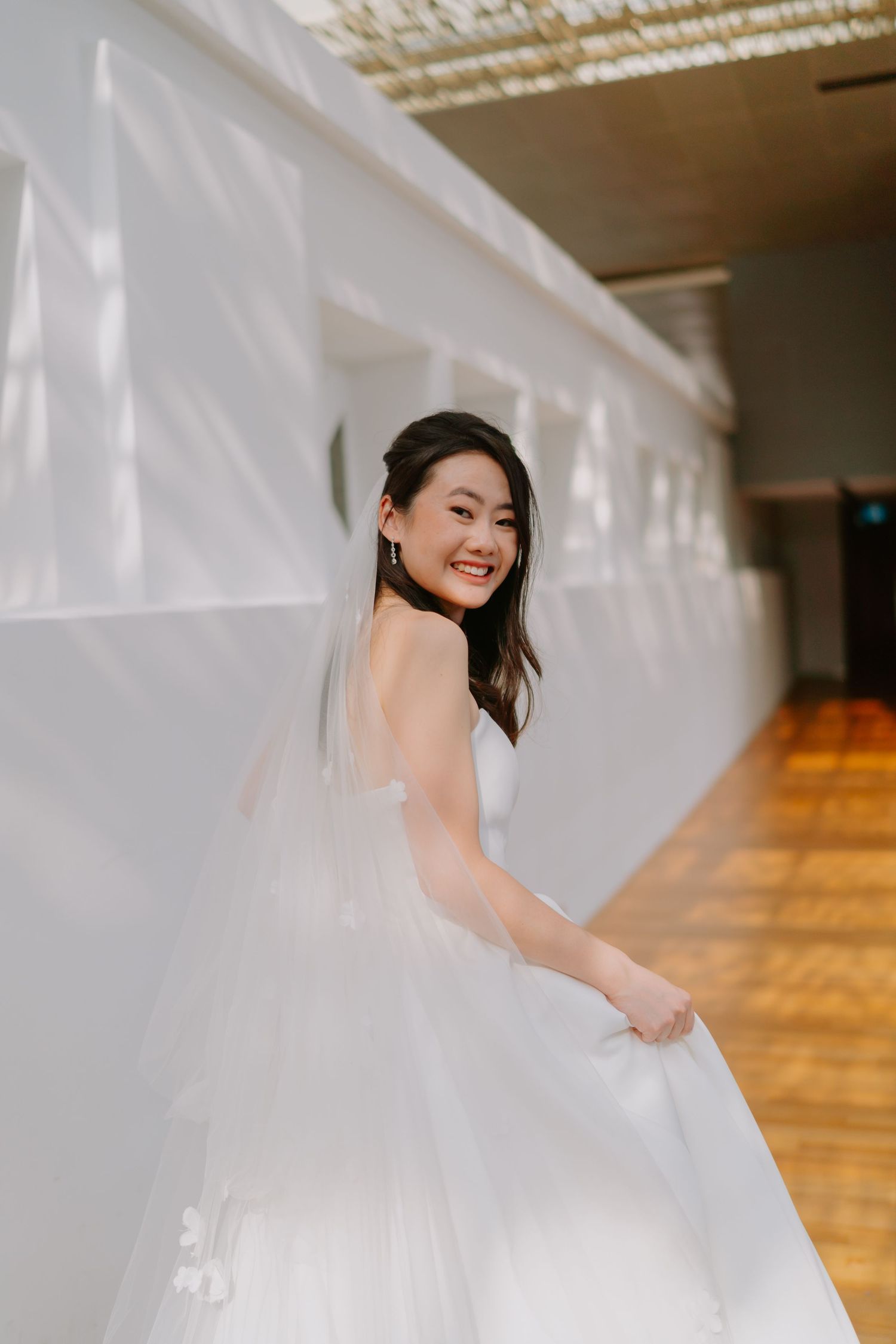 Elegant Asian bride in white ball gown smiles while posing against white wall indoors.